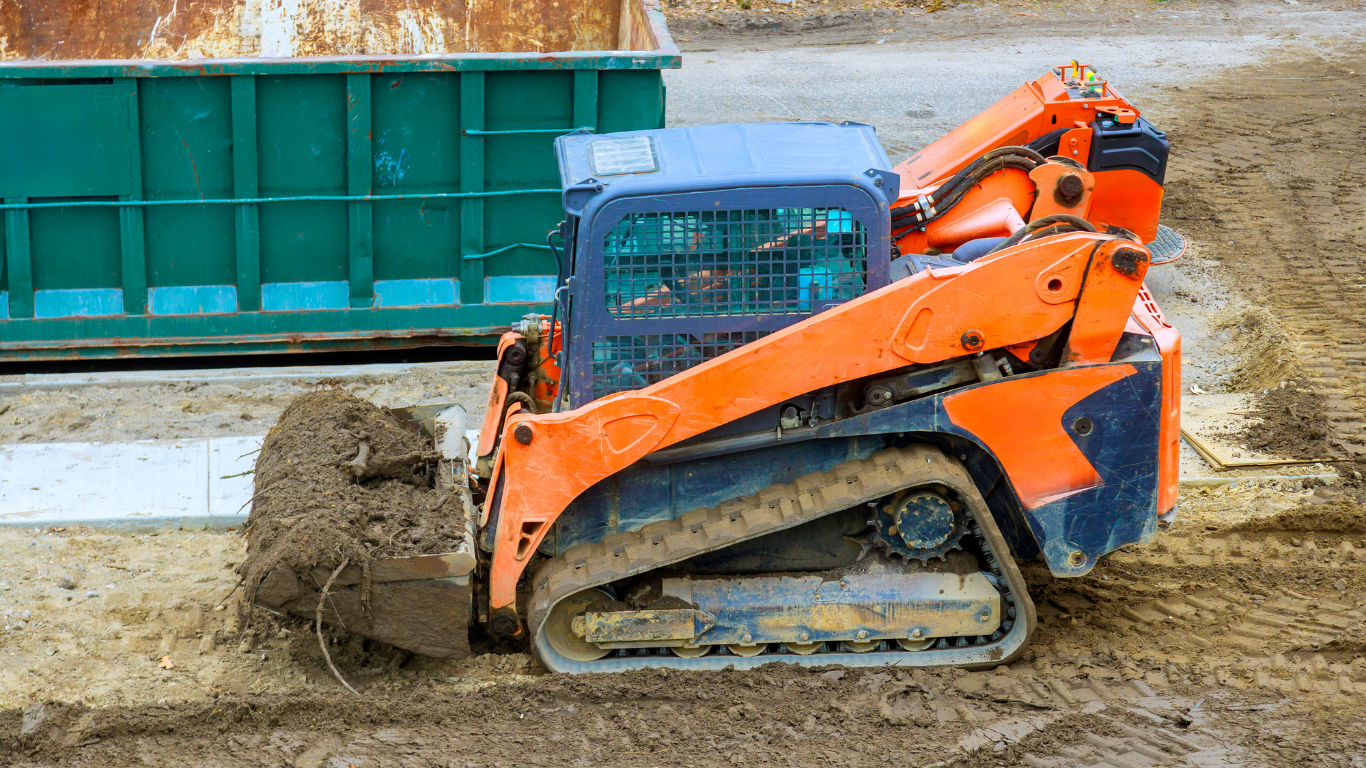 Orange skid steer loader with dirt in the bucket, tracks on muddy ground, near a green dumpster.