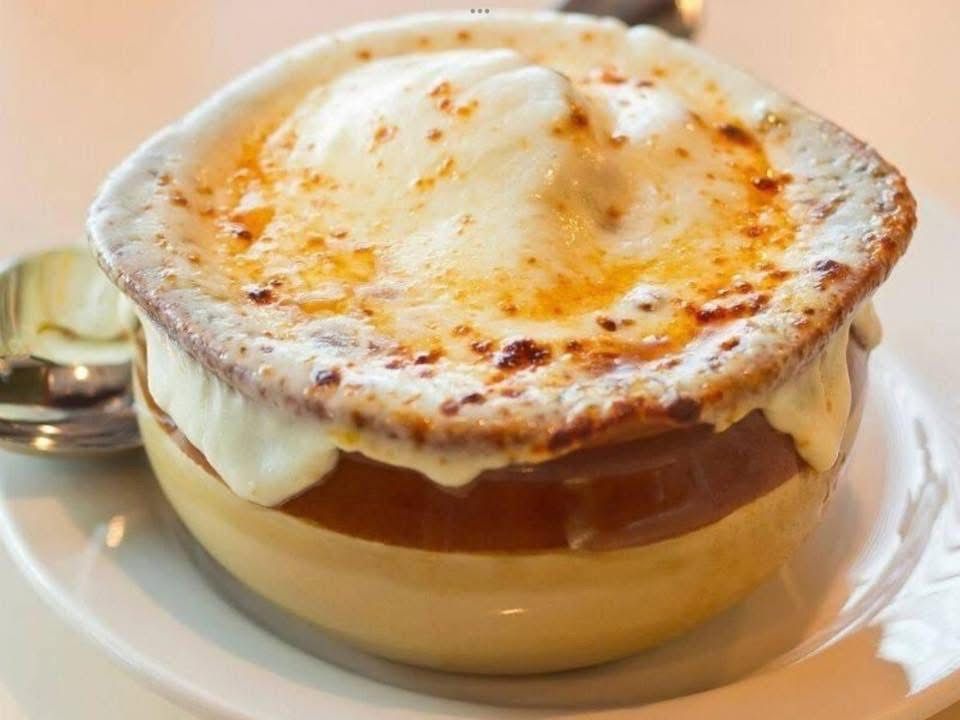 A bowl of french onion soup with cheese on top is on a plate with a spoon.