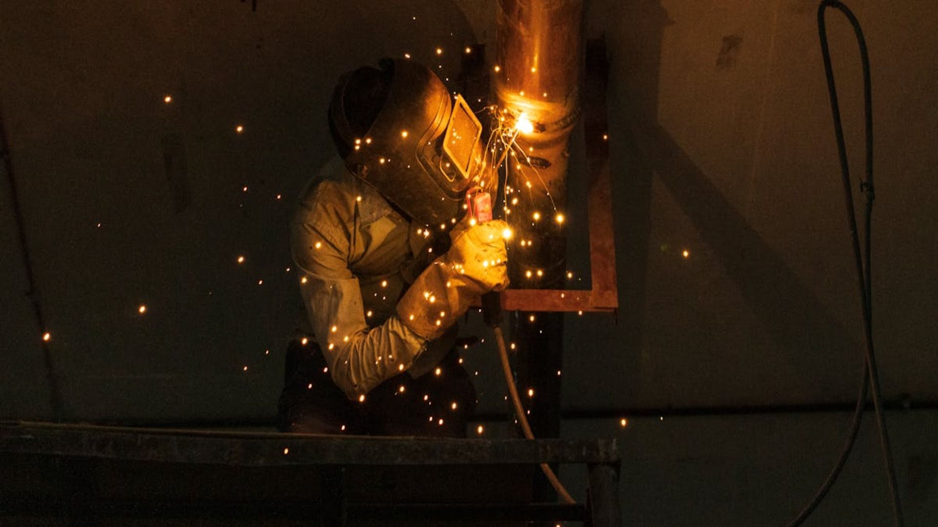 Soldador con equipo de protección trabajando en un tubo de metal, mientras saltan chispas en un taller poco iluminado.