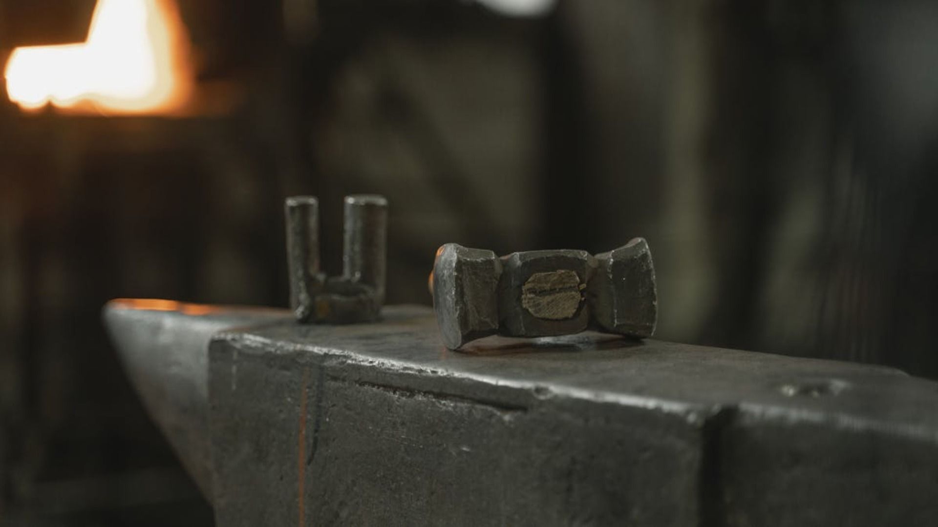 Dos piezas de metal sobre un yunque en una forja, una en forma de U, la otra un pequeño martillo; fondo ardiente.