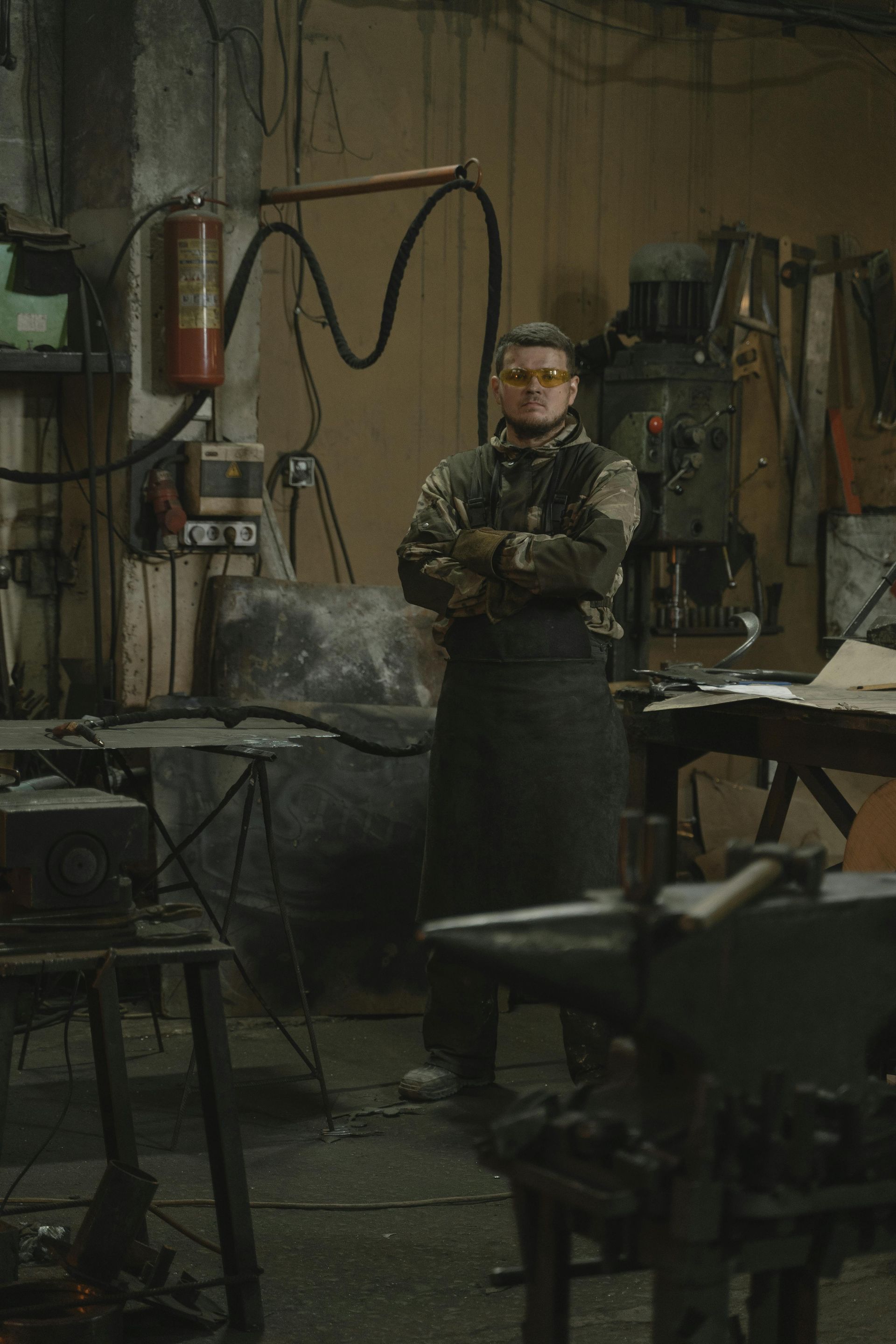 Un herrero en un taller, con los brazos cruzados, gafas y delantal.