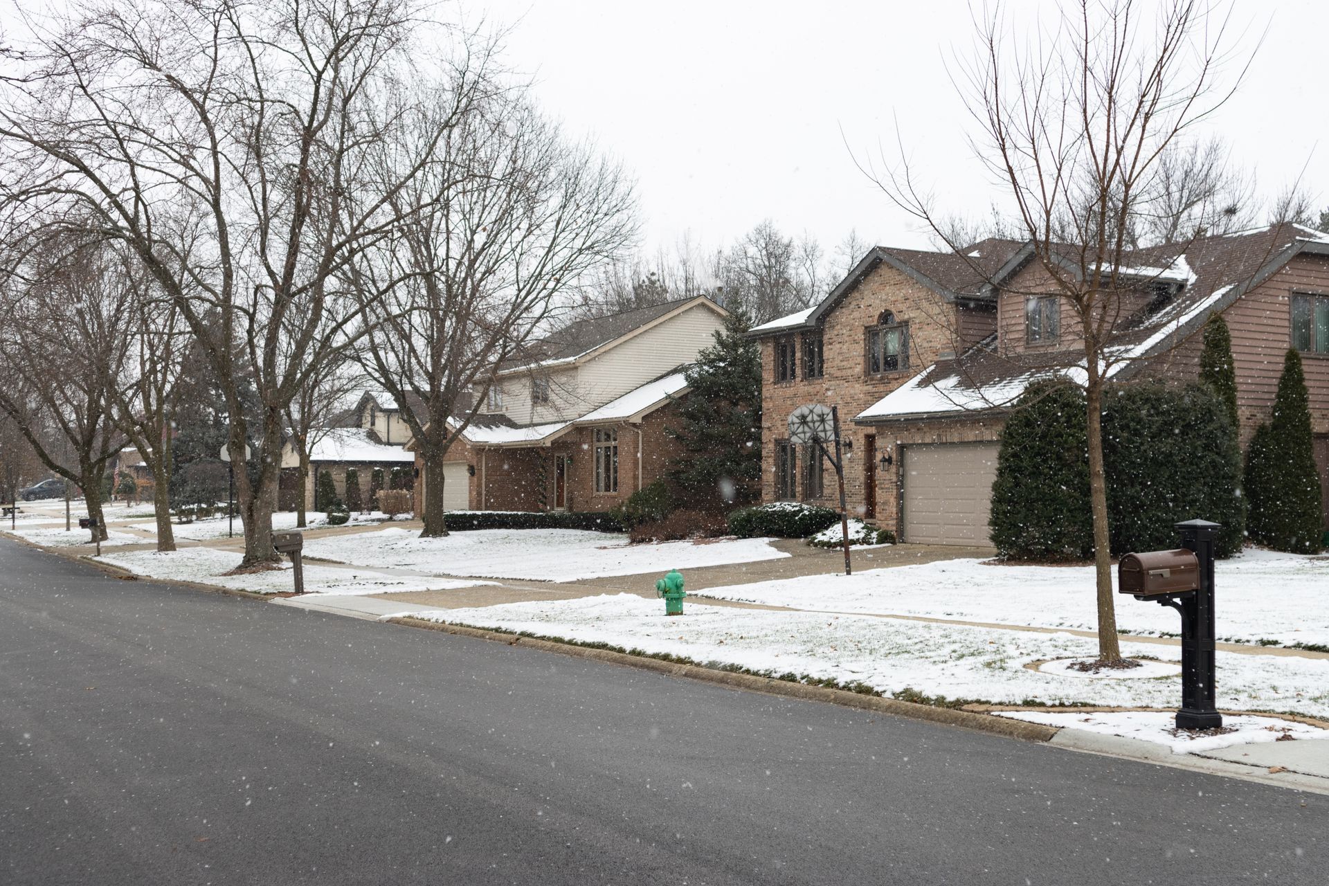 Snowy suburban street with houses, trees, and a mailbox.