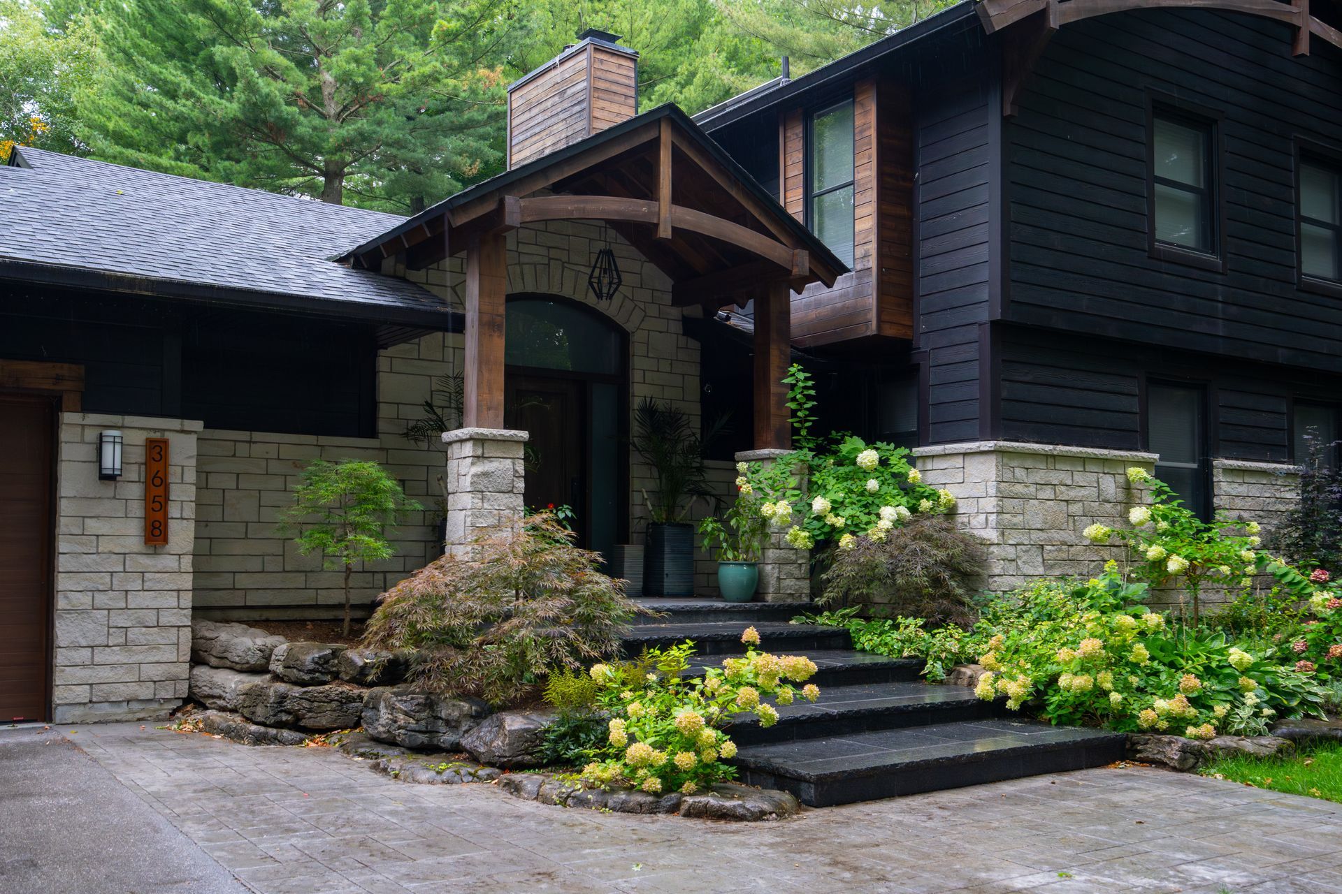 Black house with stone accents, steps, and a covered entrance surrounded by lush landscaping.