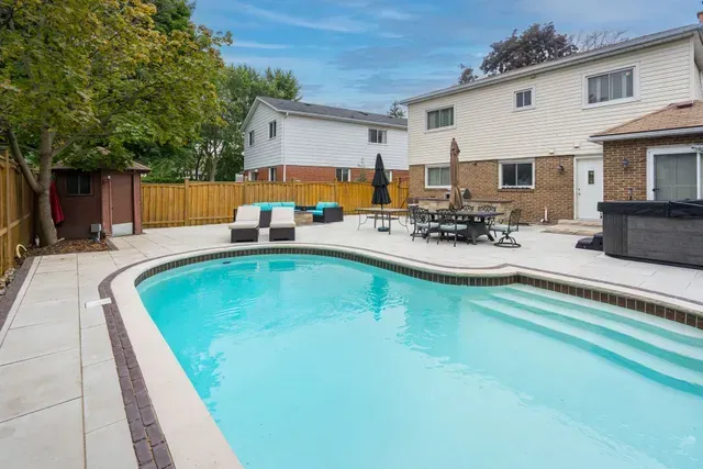 Backyard with a swimming pool, patio furniture, and two-story houses under a cloudy blue sky.