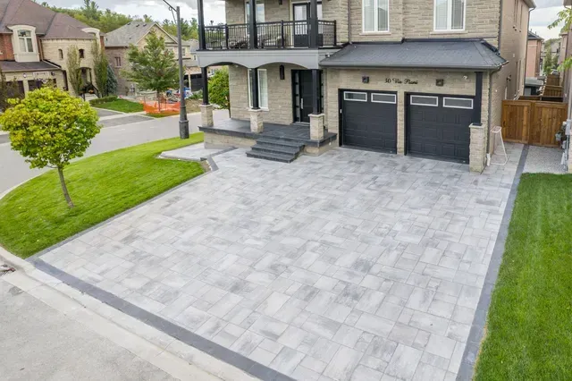 Gray paver driveway and lawn in front of a two-story home with black garage doors and balcony.