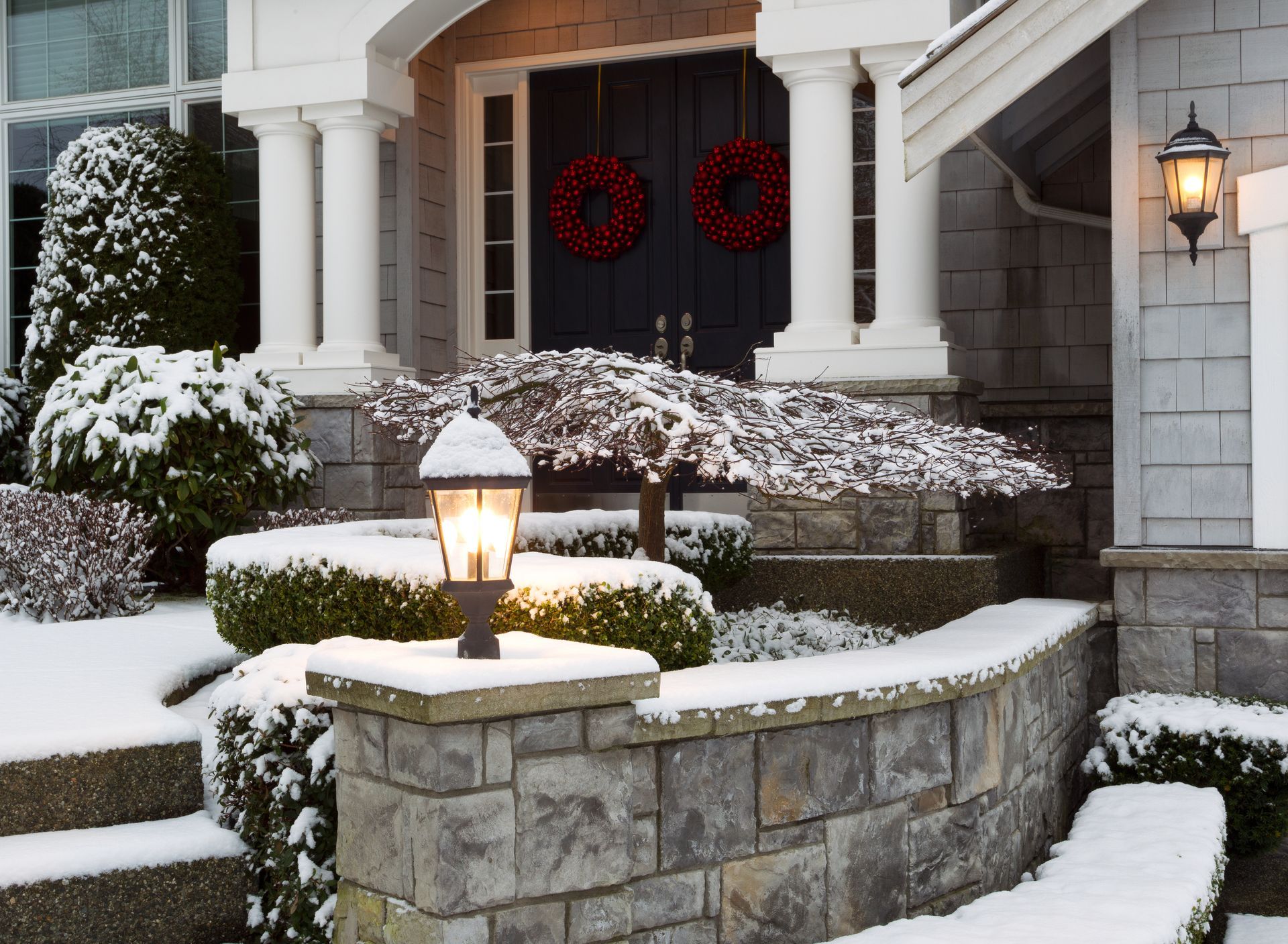 Snowy house entrance with stone wall, lamp post, and wreaths on the door.