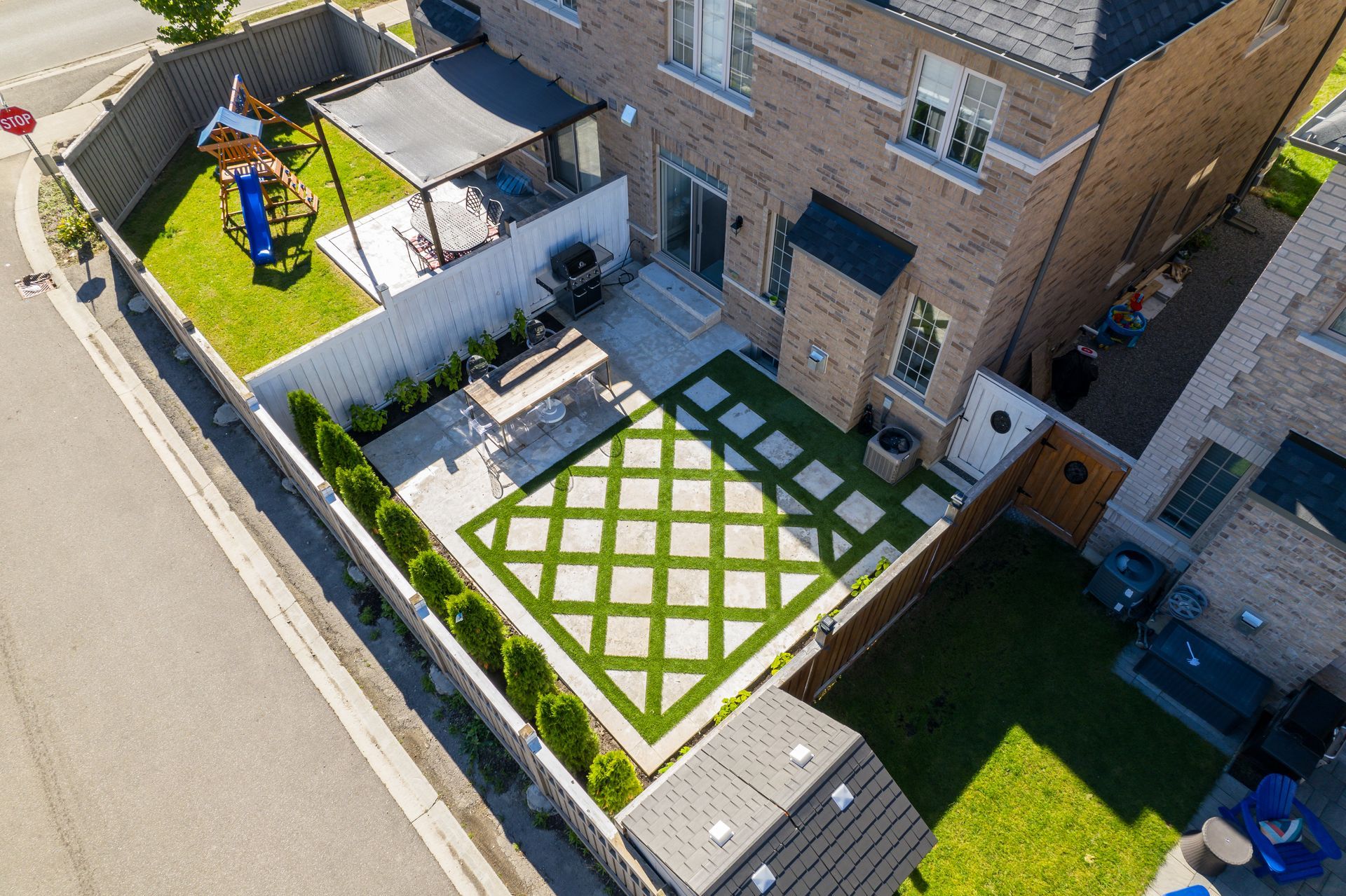 Aerial view of a backyard with a patio, lawn, and play structure, surrounded by a brick house and fences.