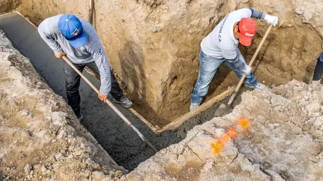 Two workers shoveling dirt in a trench, one in a blue cap, the other in a red cap.