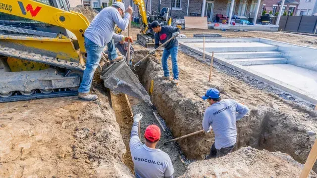 Construction workers pouring concrete into a trench near a pool. A yellow excavator is visible.