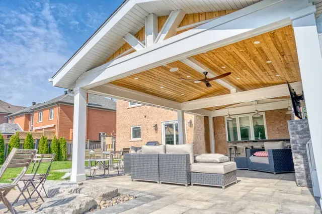 Outdoor patio with seating under a white-framed wooden ceiling. View of a brick house and blue sky.