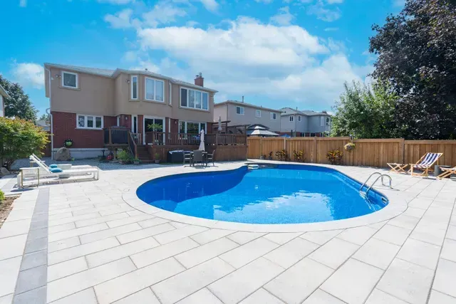Backyard with blue swimming pool surrounded by stone patio, house in background, sunny day.