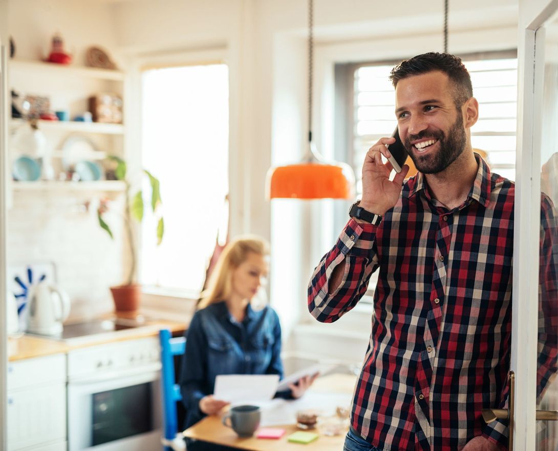 A man is talking on a cell phone in a kitchen while a woman sits at a table.