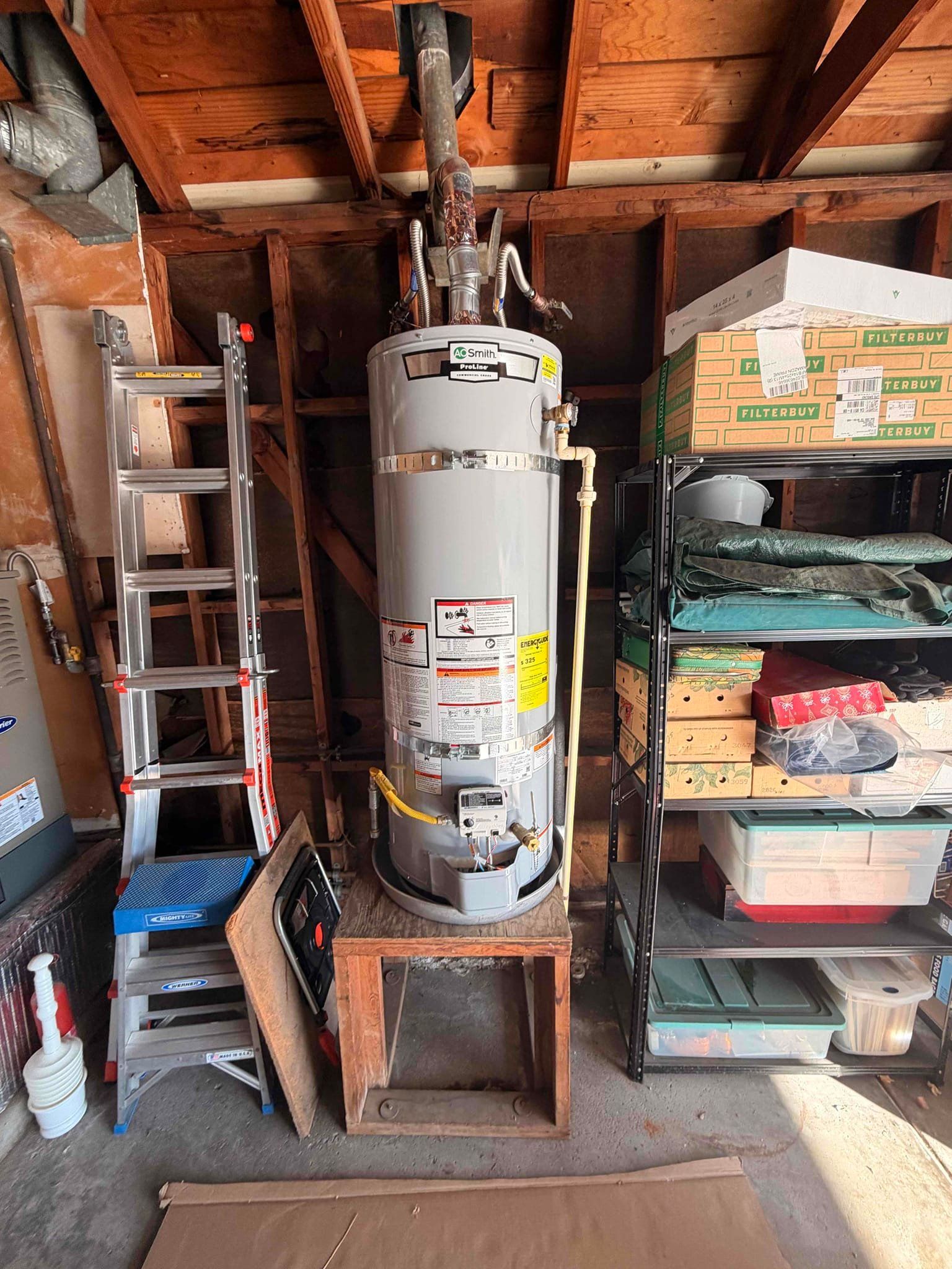 A tall, gray water heater stands in a dark, unfinished basement with exposed wooden ceiling joists and pink insulation.