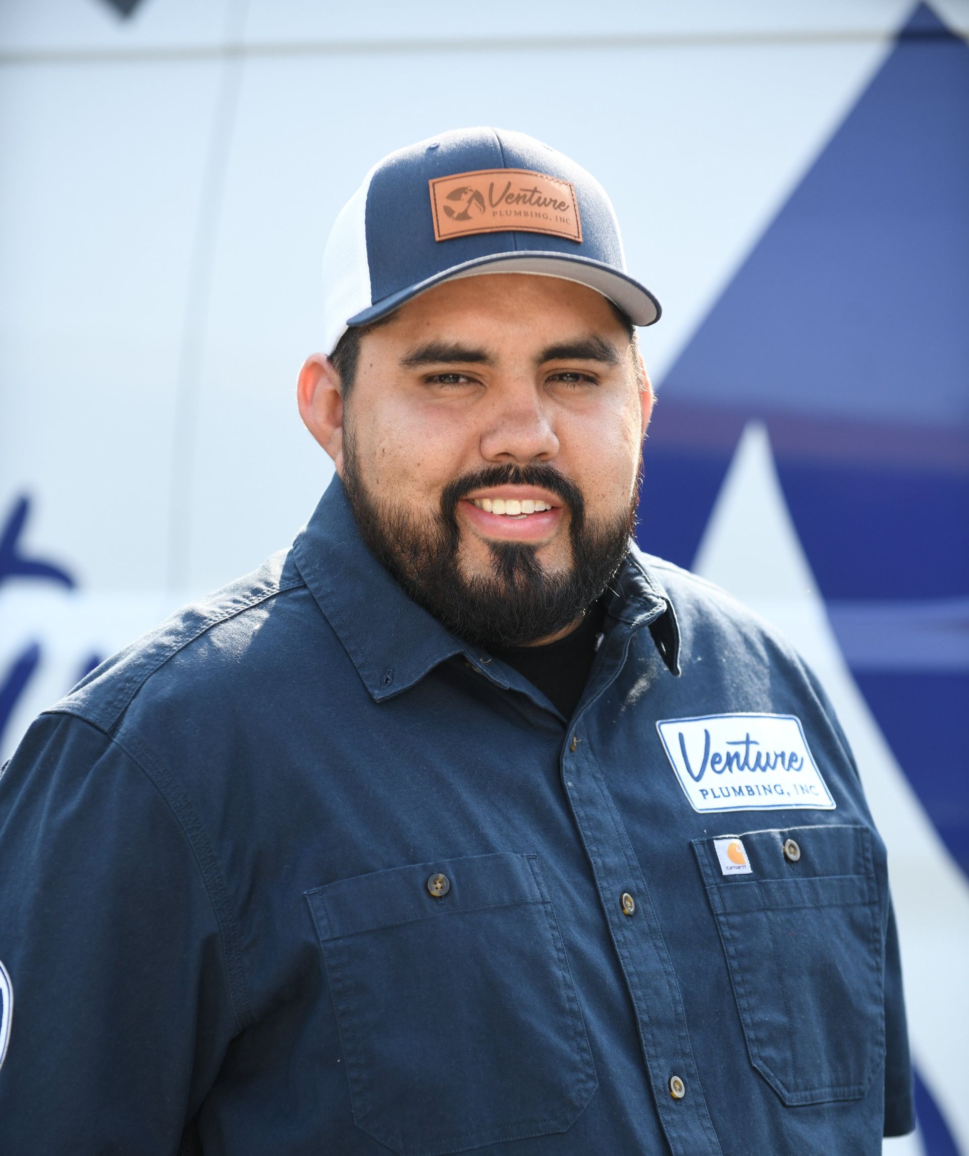 A person in a blue work shirt and cap with a logo stands in front of a blue and white vehicle, smiling at the camera.
