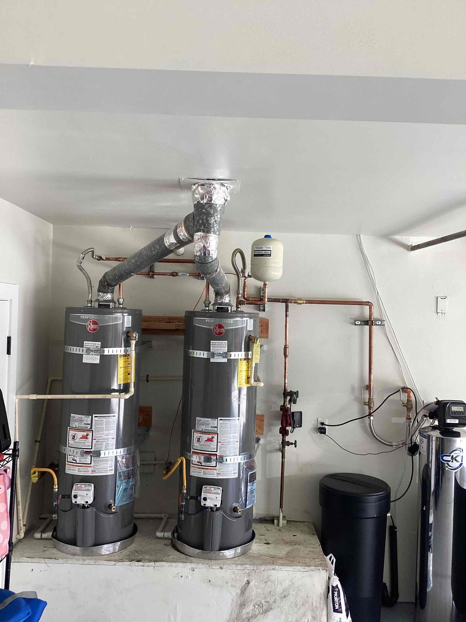 Two gray residential water heaters stand on a concrete base with a shared vent pipe and connected plumbing in a garage.