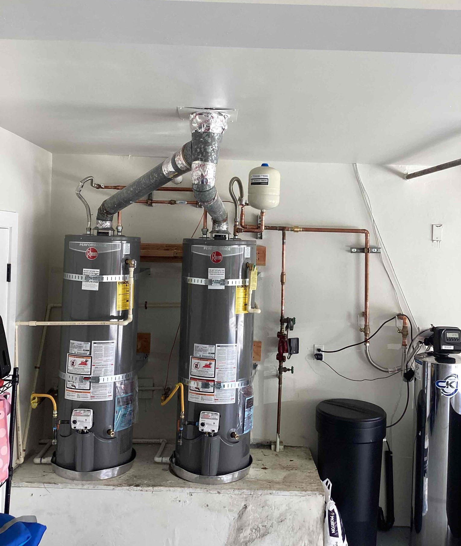Two gray water heaters connected to a shared ceiling exhaust vent, sitting on a concrete platform in a utility room.