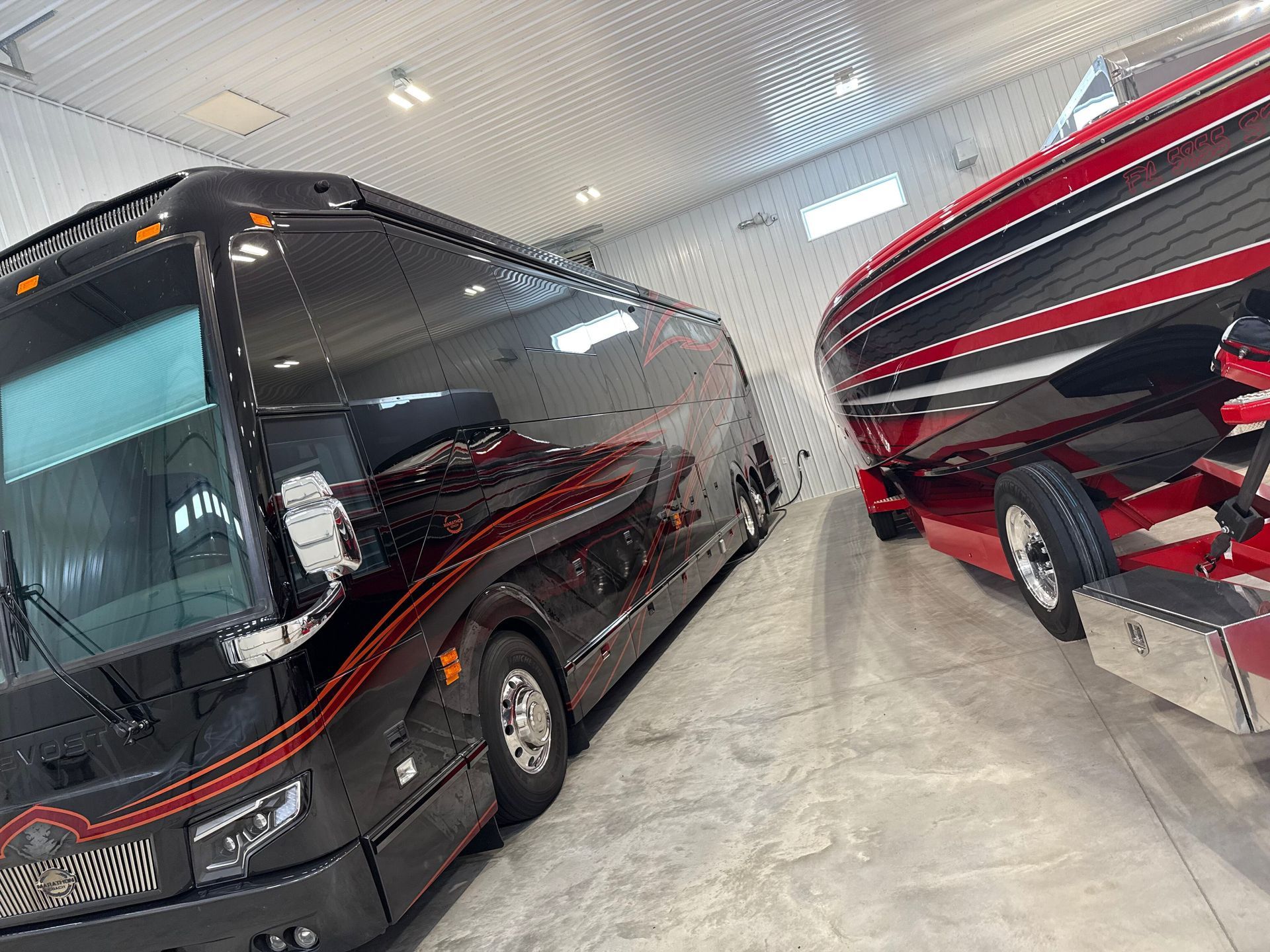 A black bus is parked next to a red boat in a garage.