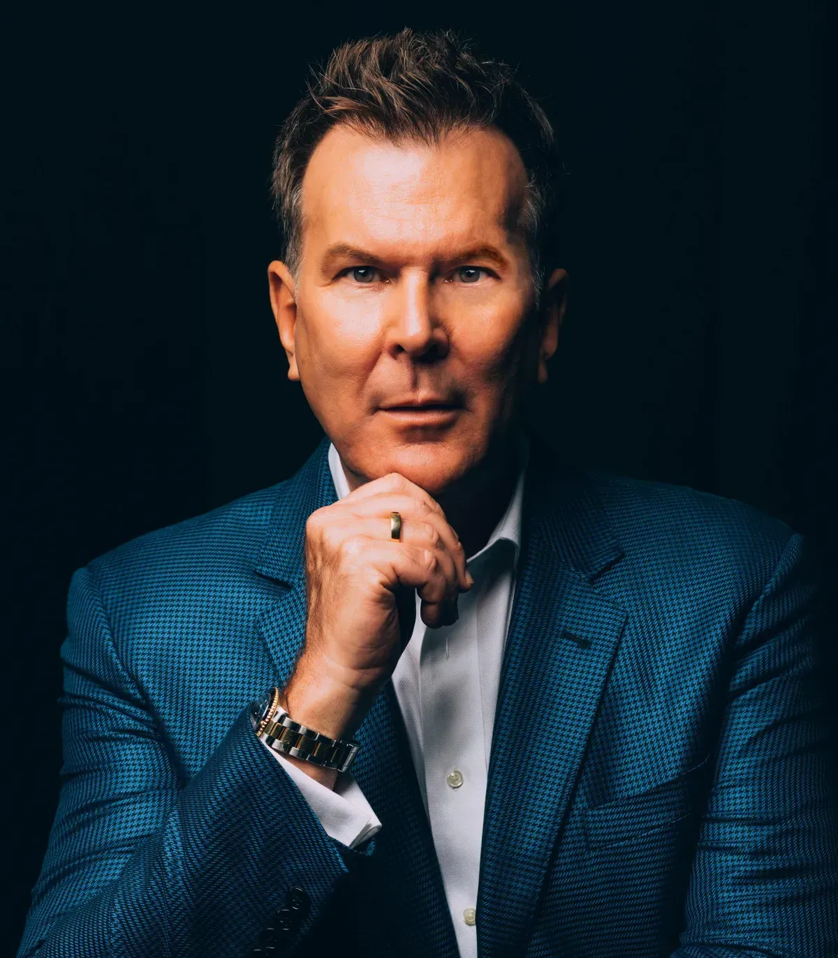 A man in a blue suit and white shirt is holding his hand to his chin.