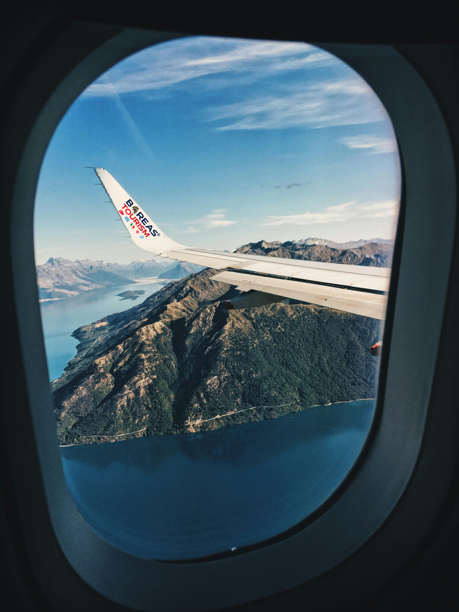 Airplane wing over mountains and water, viewed from a plane window on a clear day.