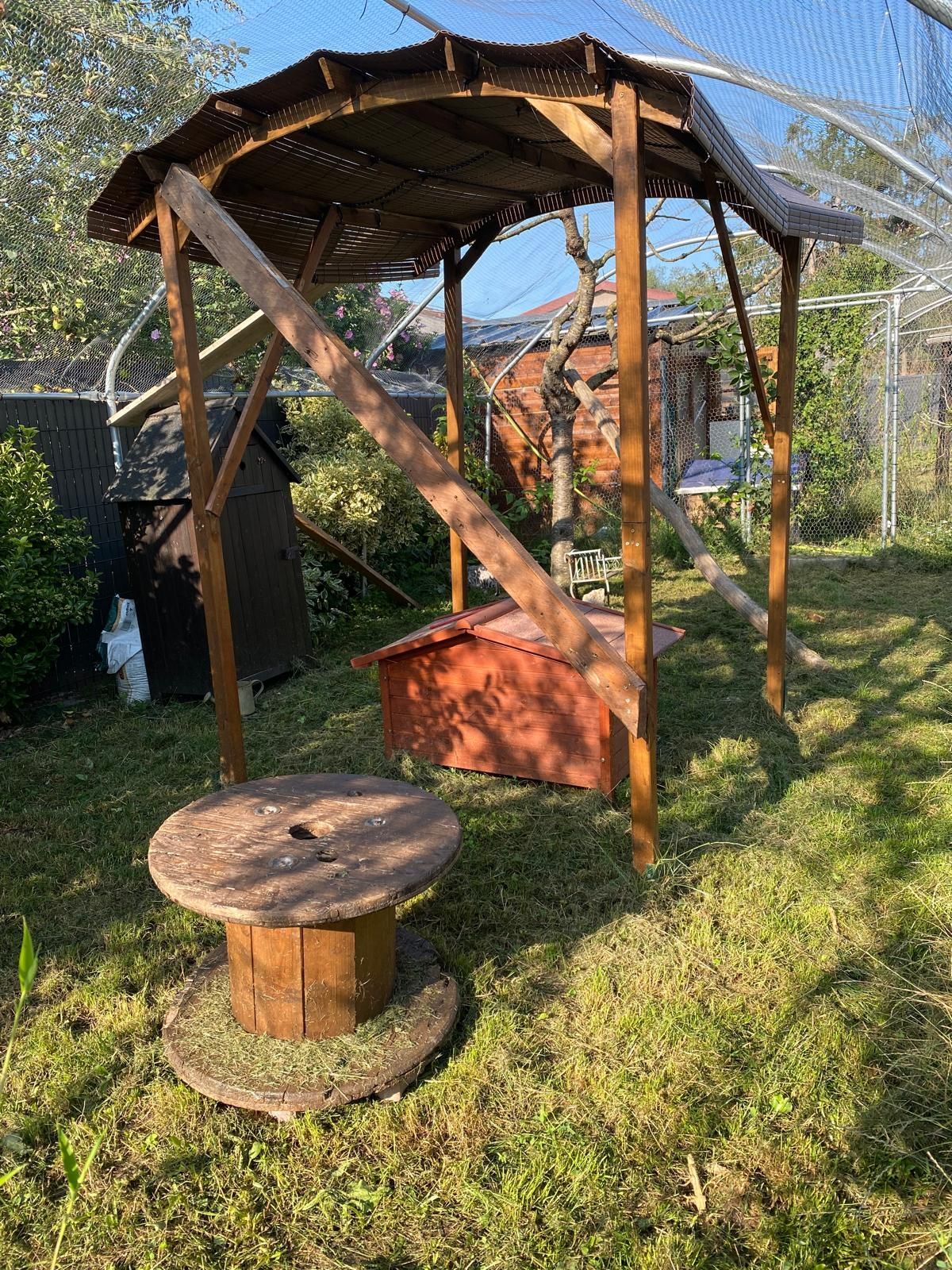 Gazebo en bois avec une table à bobines et une boîte rouge dans une cour herbeuse.
