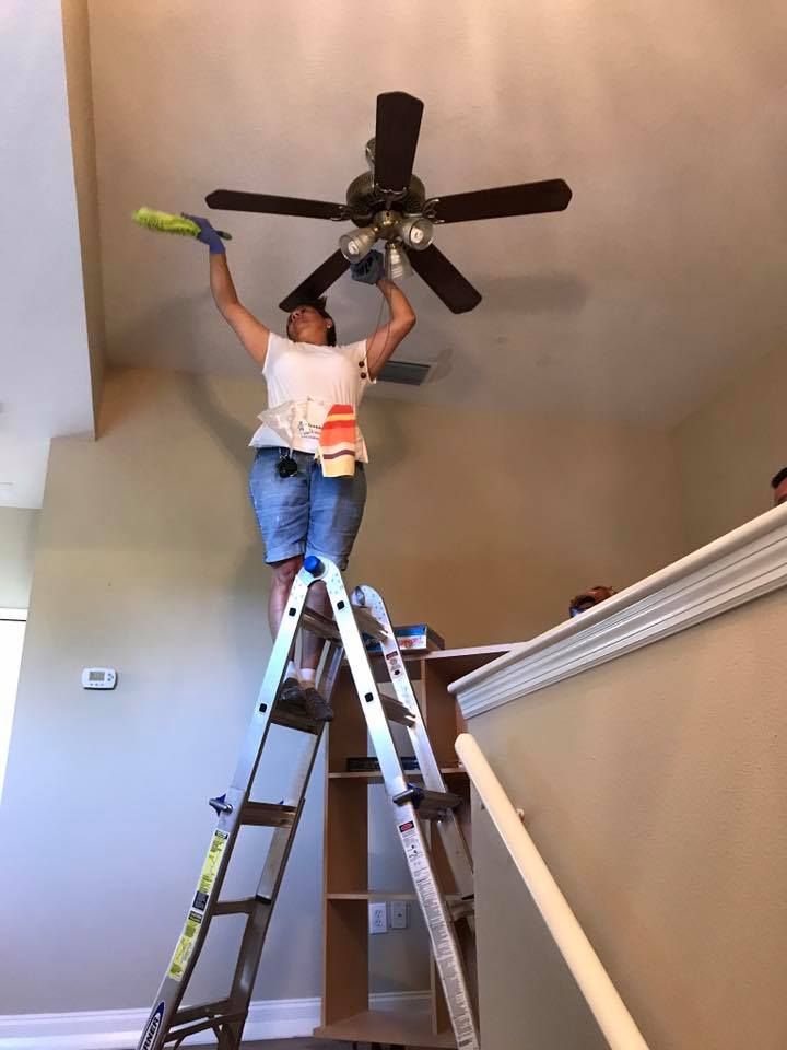 A woman is standing on a ladder cleaning a ceiling fan.