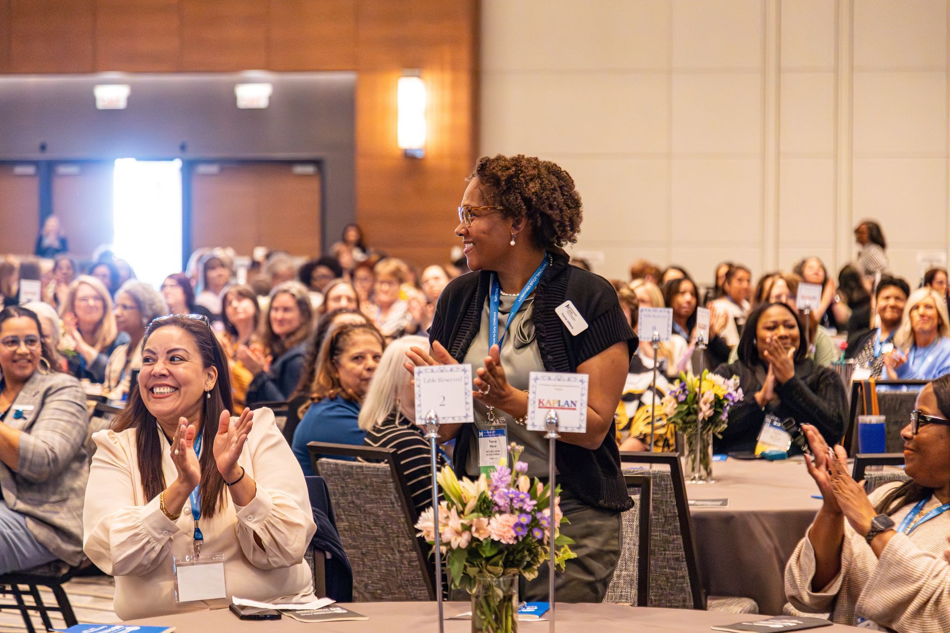 A crowd of people seated at round tables in a conference hall are clapping, with some individuals standing to cheer.