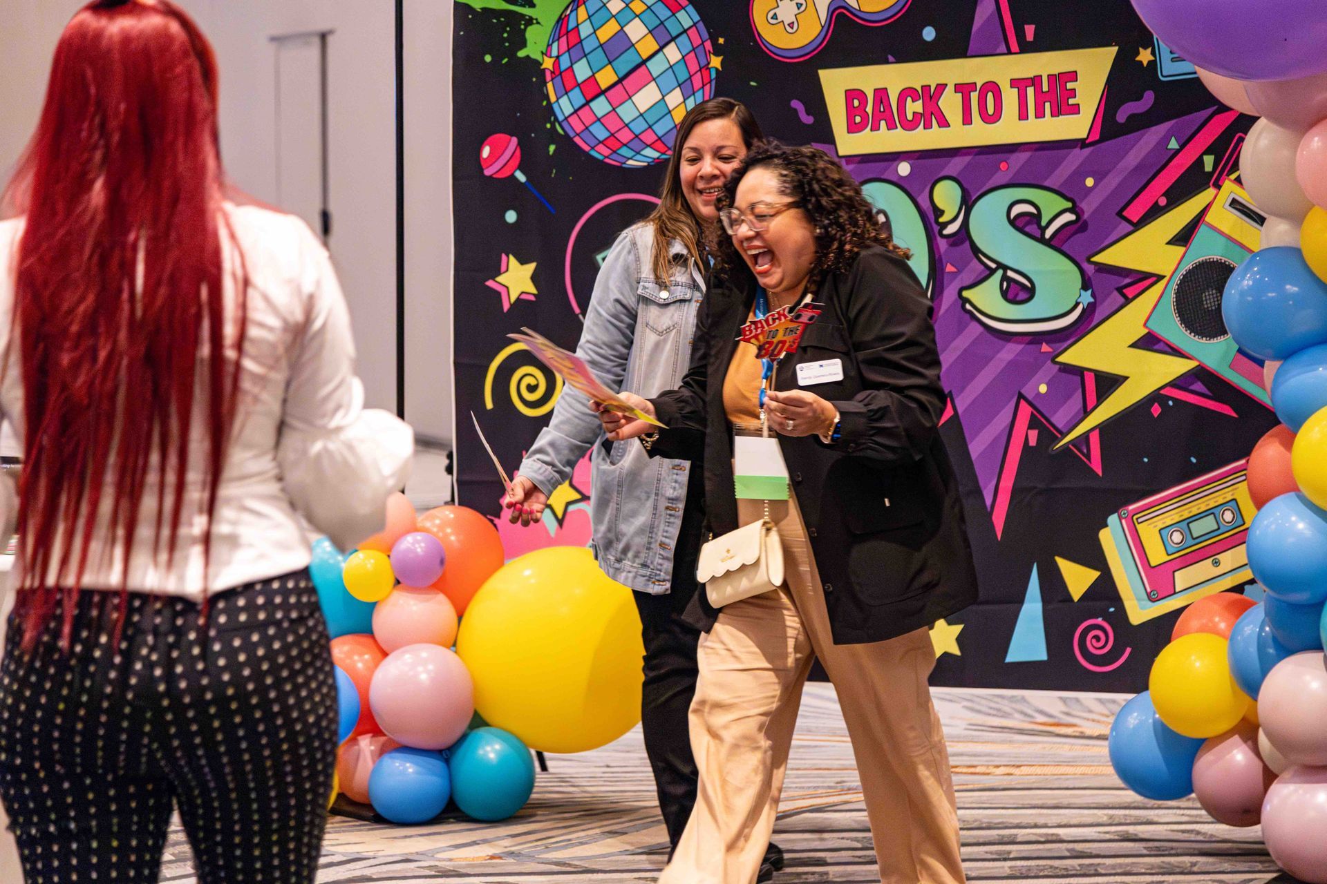 Two women are standing in front of a back to the 80 's backdrop.