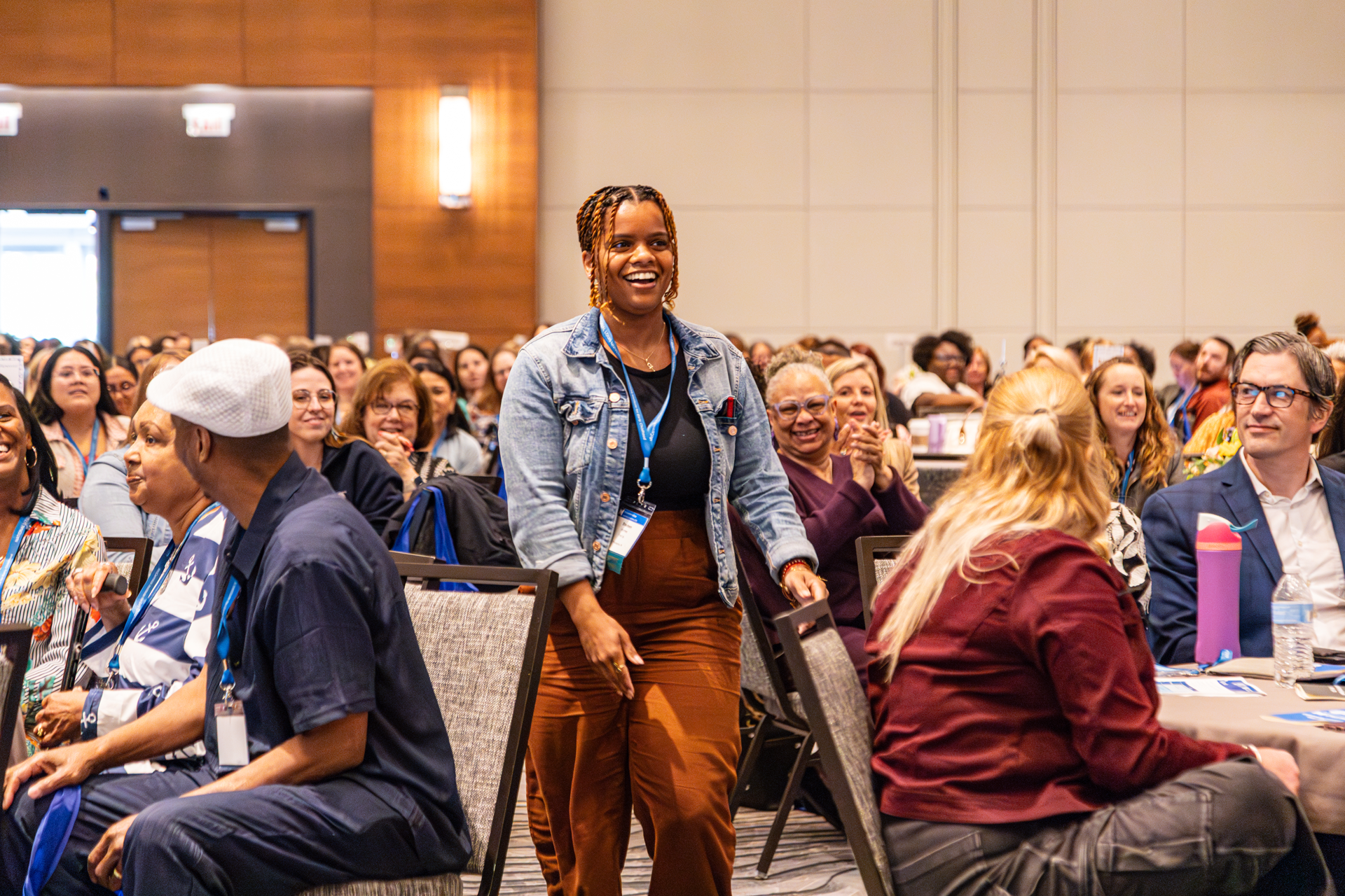 Woman in denim jacket walks through seated audience in large conference room.