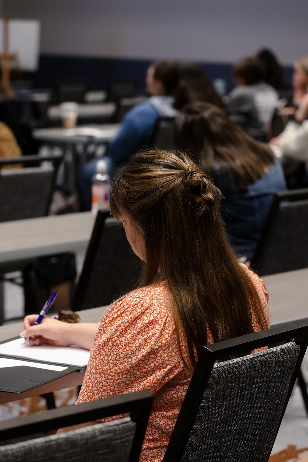 A woman is sitting at a table in a classroom writing in a notebook.