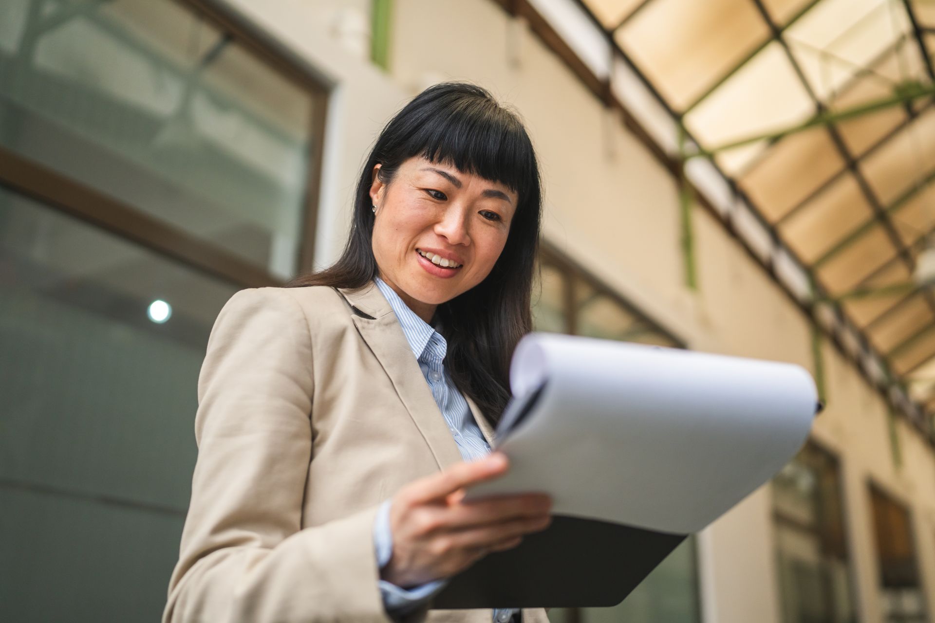 Woman in a blazer, reading papers on a clipboard, smiling. Indoors, natural light.