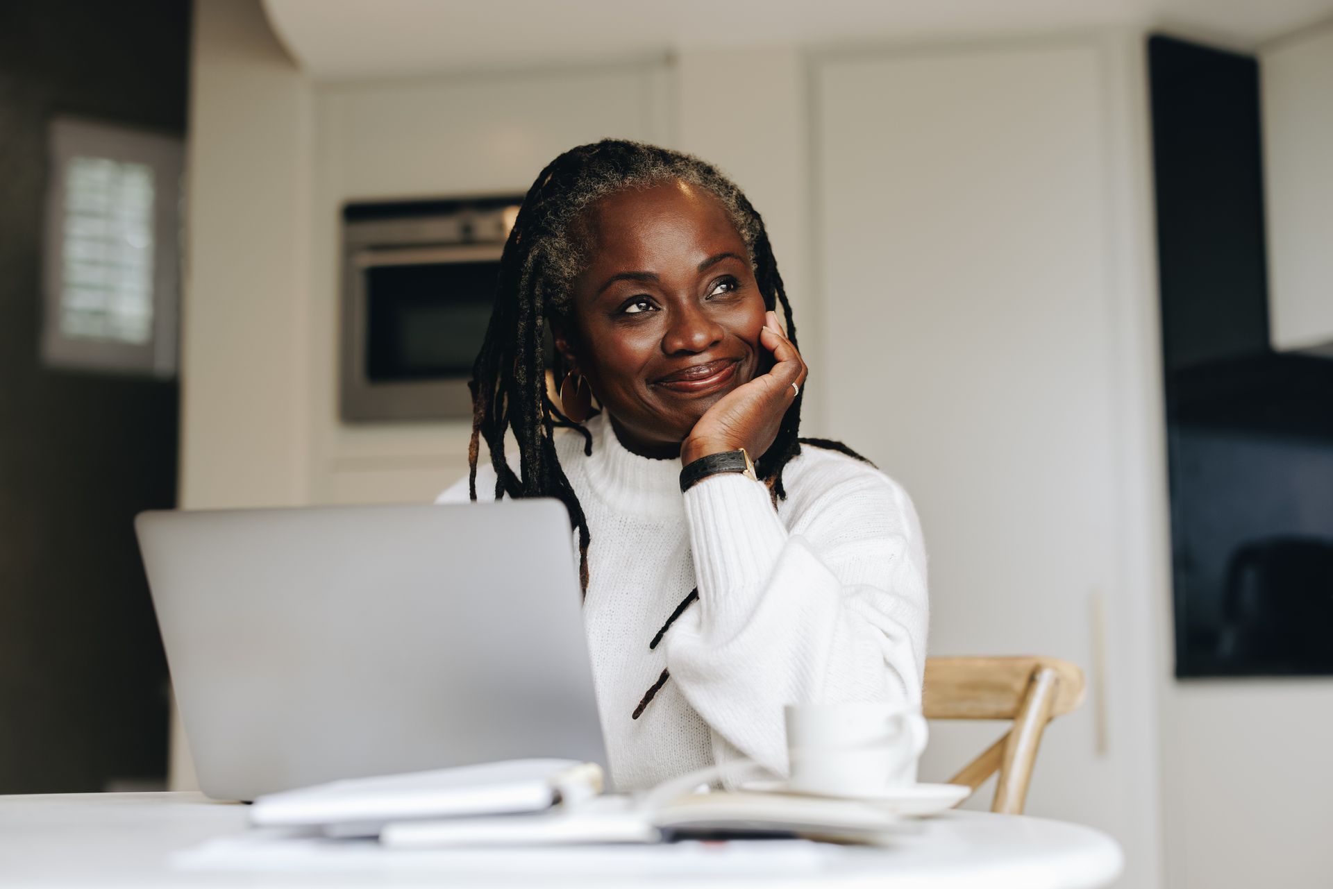 Woman with dreadlocks, smiling, resting her chin, looking up, near a laptop, coffee, and papers.