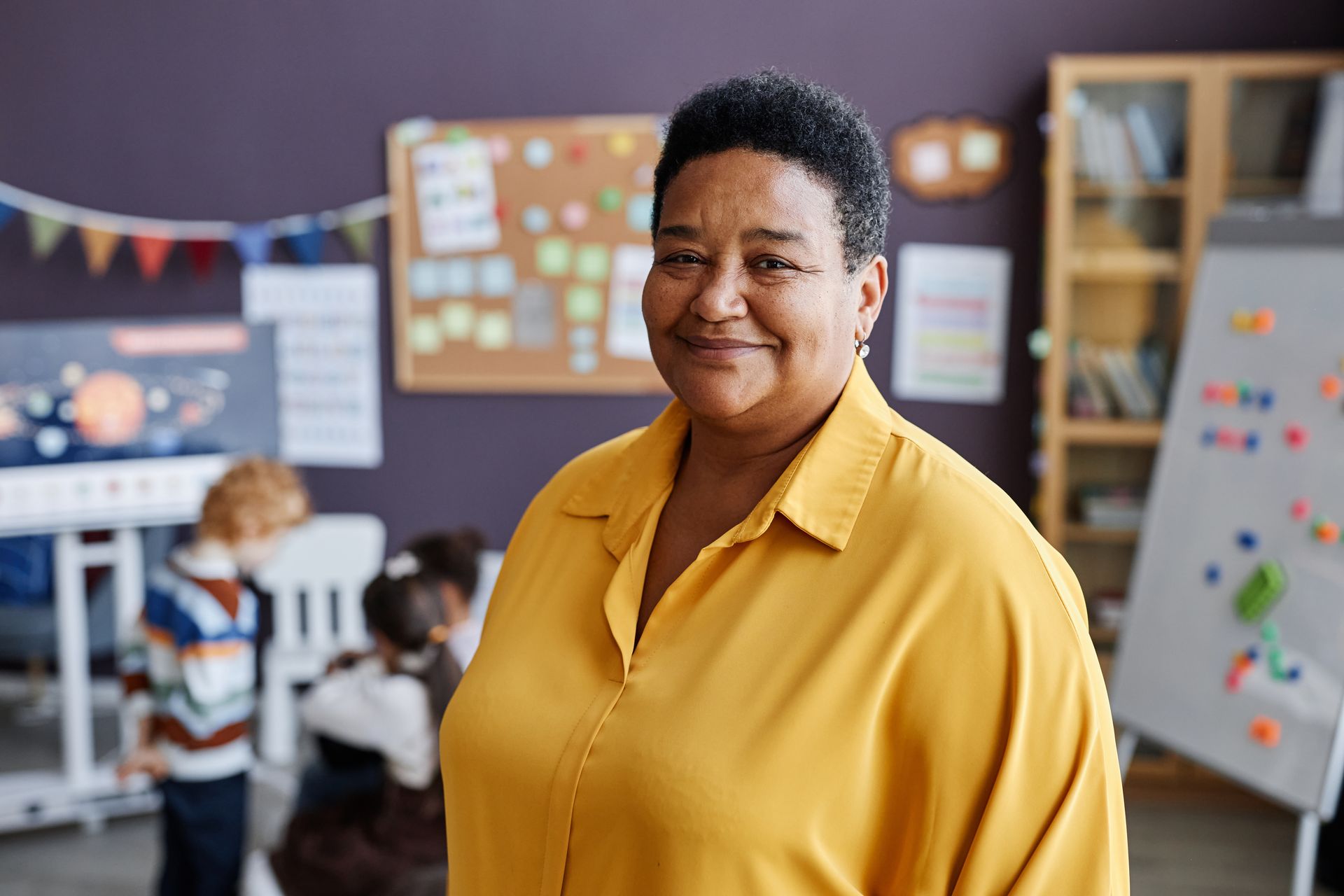 Smiling Black woman in yellow shirt stands in a classroom with children in the background.