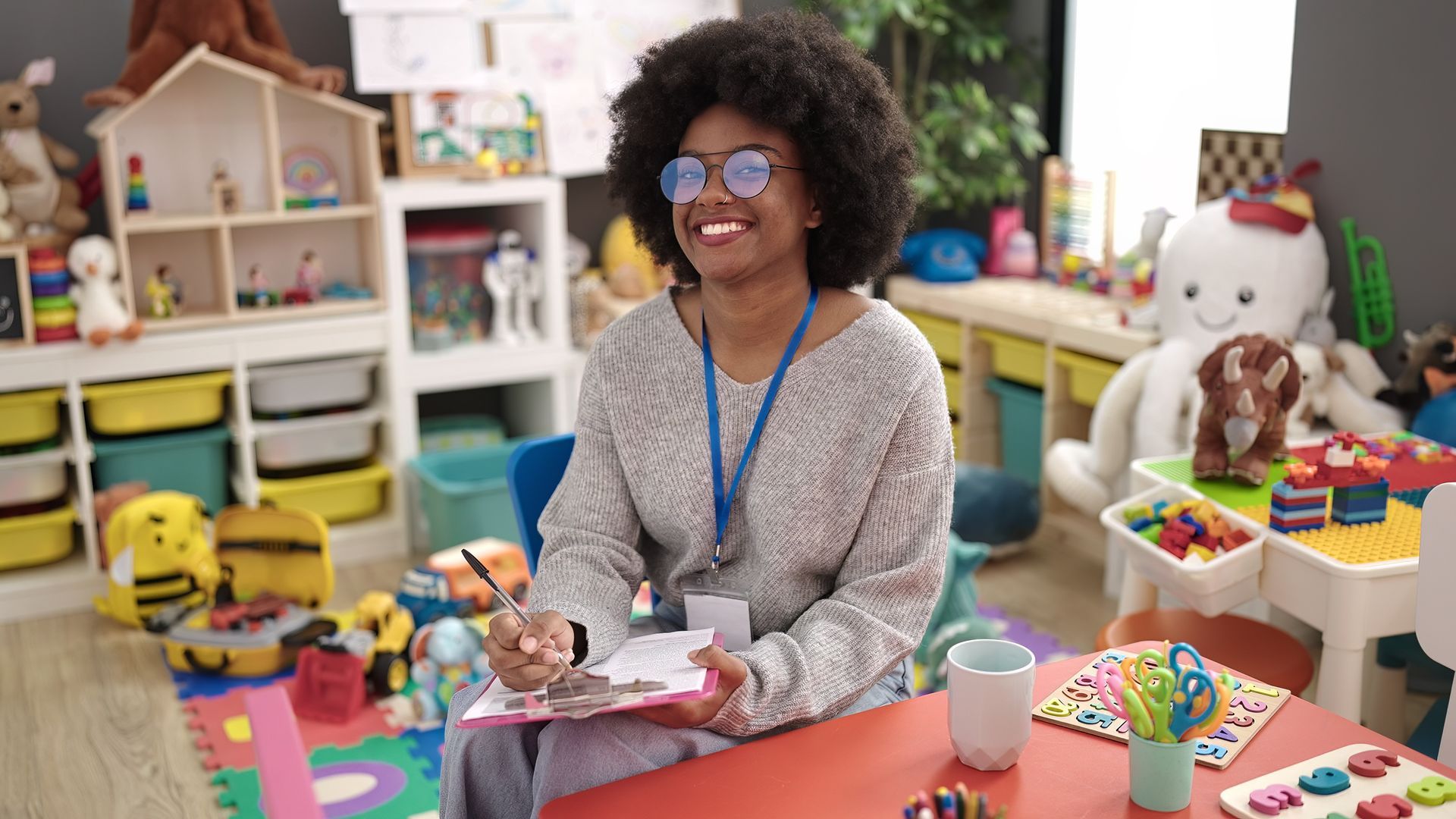Smiling Black woman in glasses writes at a table in a colorful classroom setting.