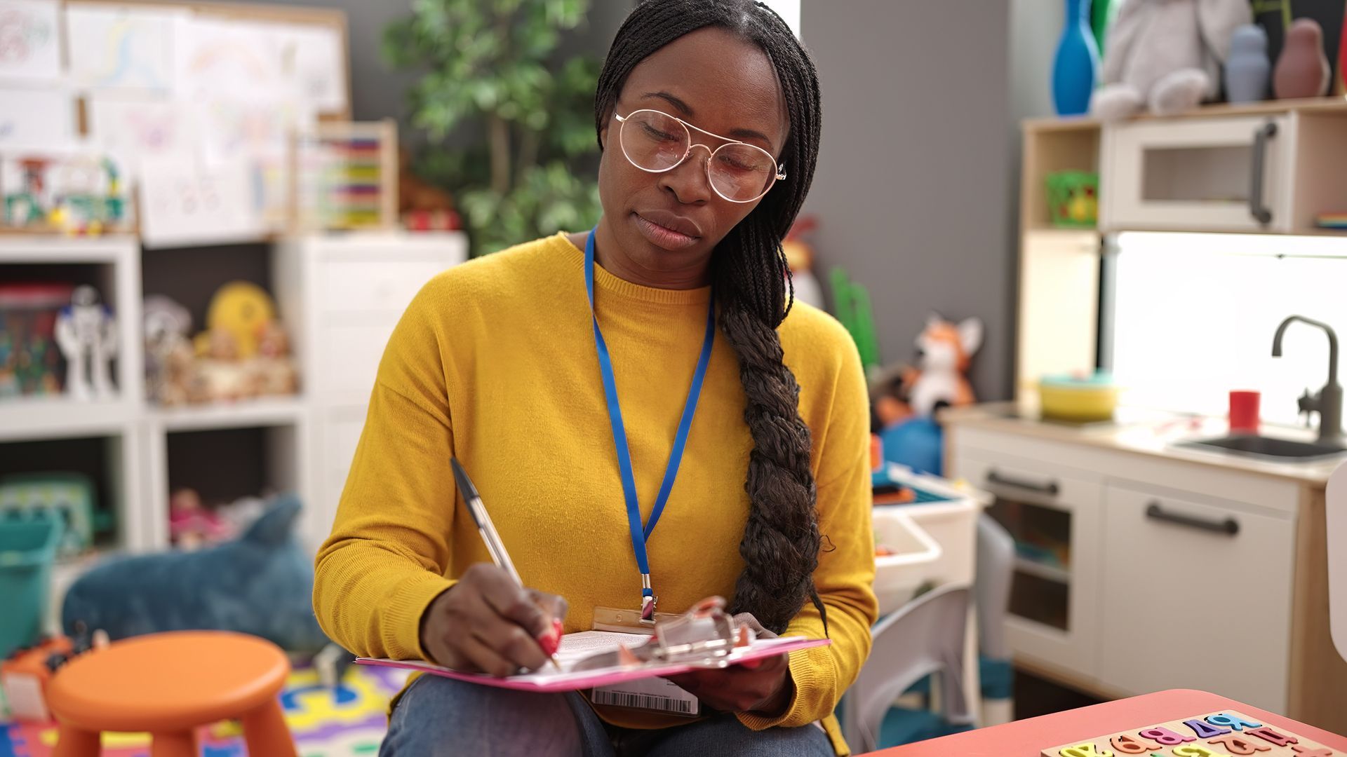 Woman in a yellow shirt writing on a clipboard in a colorful classroom.