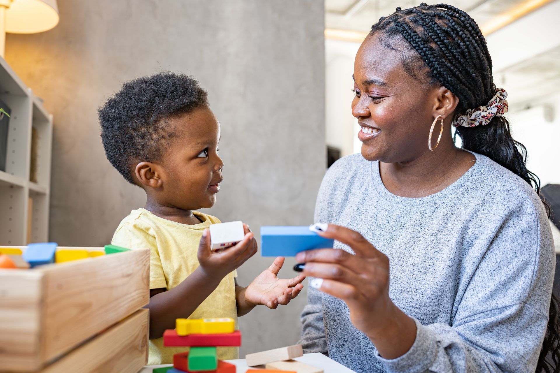 Woman and young child playing with colorful wooden blocks indoors. The woman smiles while holding a blue block.