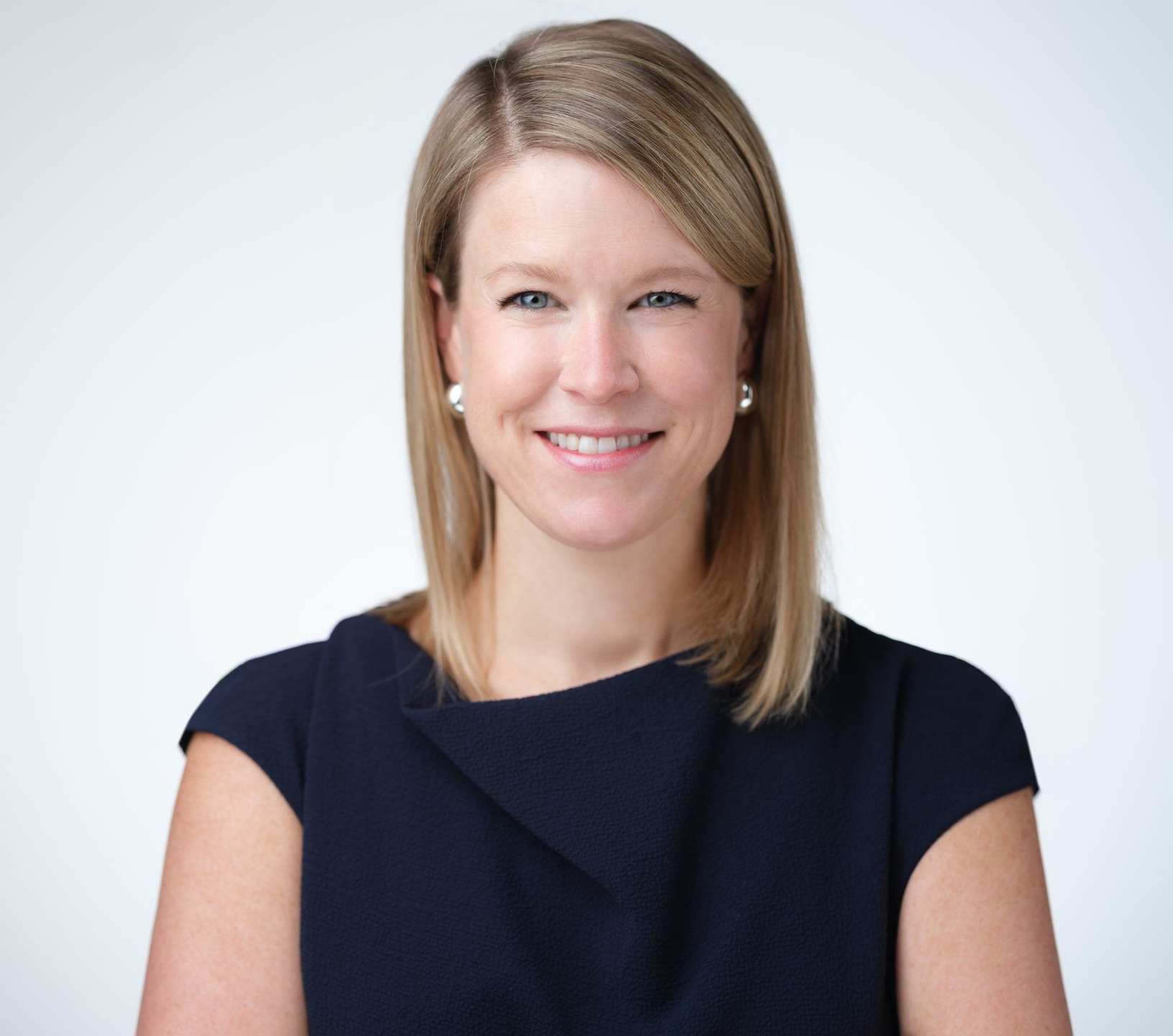 A professional headshot of a person with shoulder-length blonde hair wearing a dark blue dress against a white background.