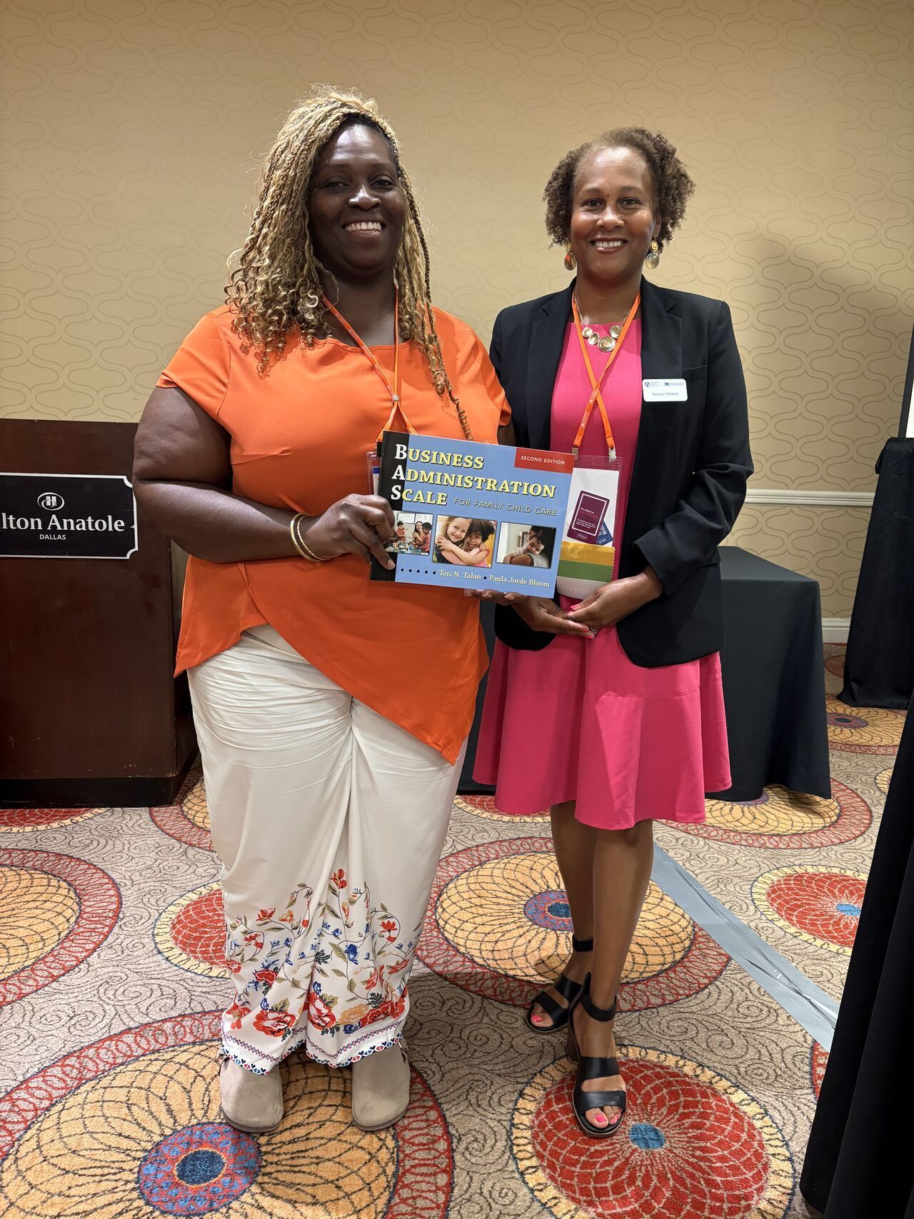 A group of women standing in front of a screen that says program administration scale Two women pose, holding a book. One wears orange, white skirt, the other a pink dress and blazer, at a conference.