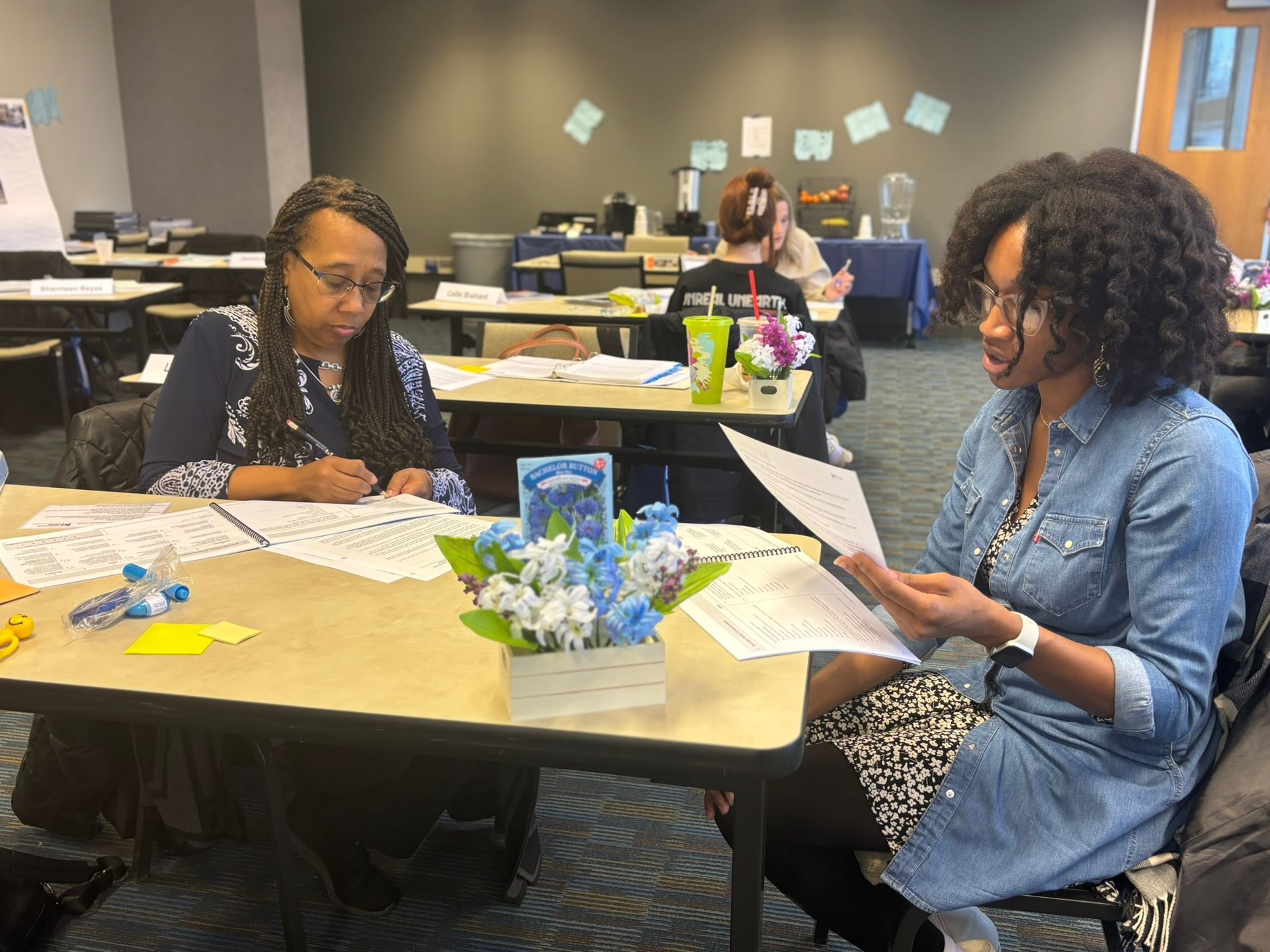 Two people seated at a table in a meeting space, one writing on a document and the other holding papers while speaking. Two people seated at a table in a meeting space, one writing on a document and the other holding papers while speaking.