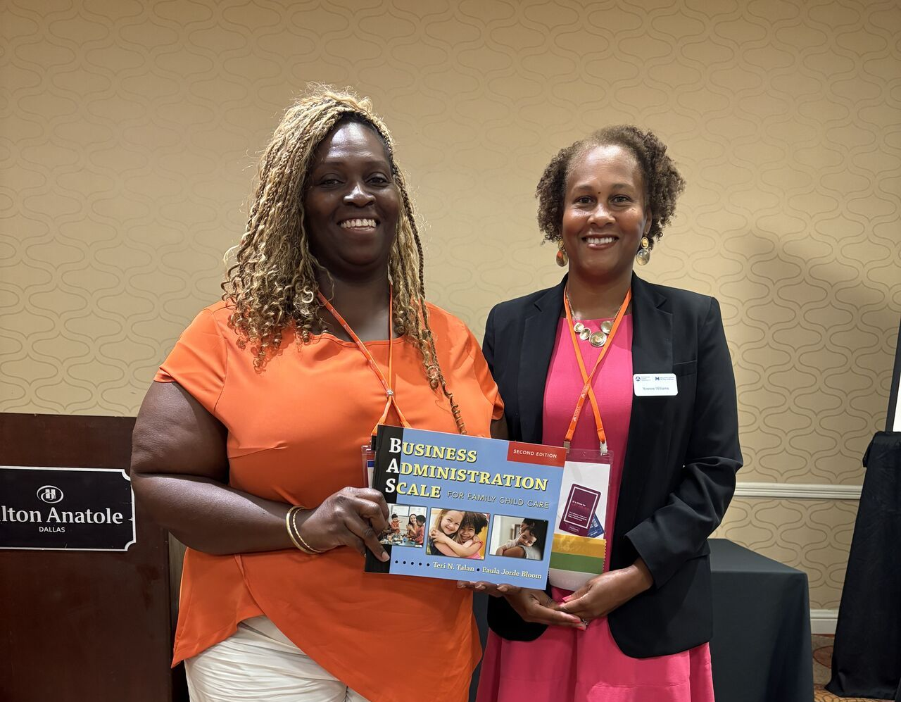 Two women smiling, holding a book. One in orange shirt, one in blazer. Standing indoors at a conference.