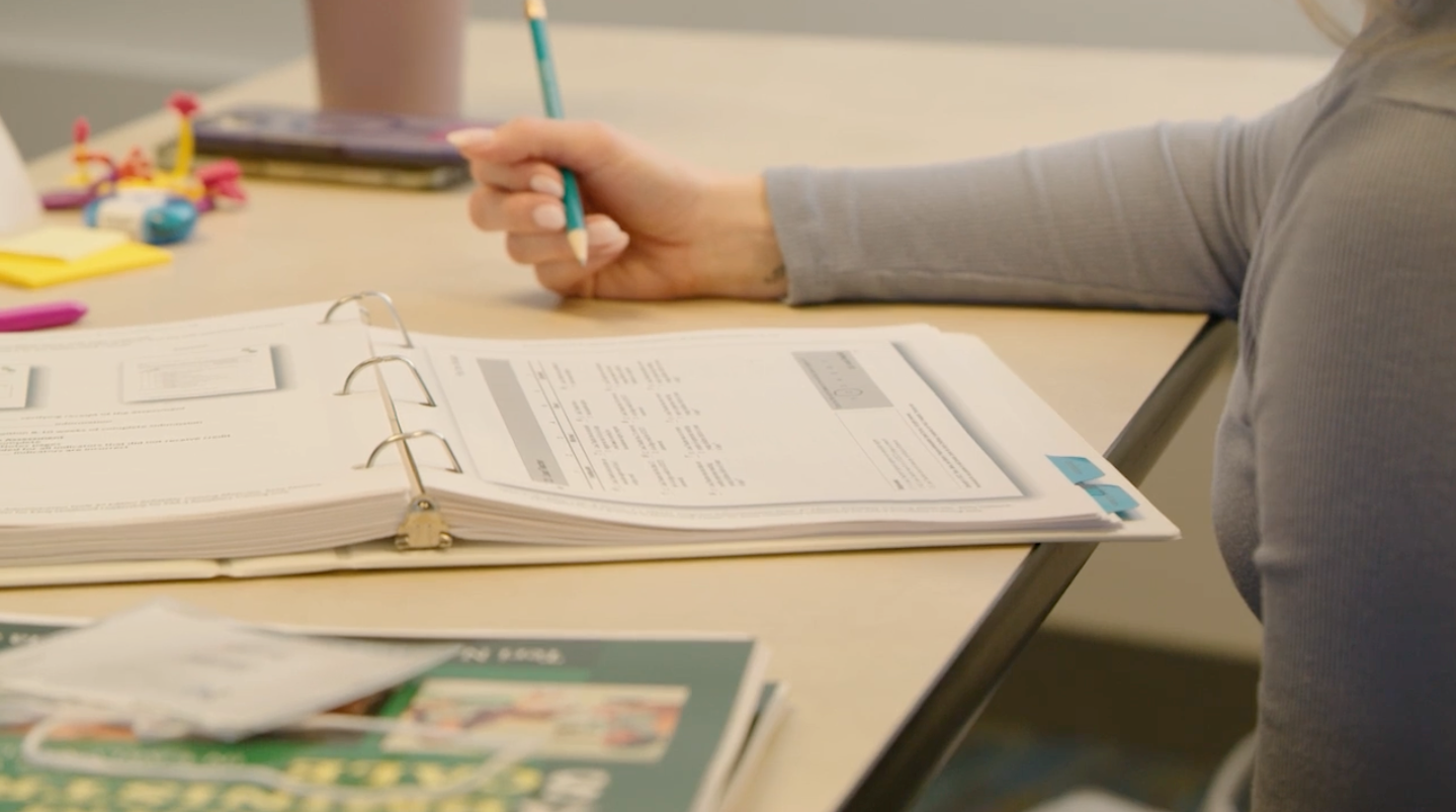 Person writing in an open binder with a pencil at a desk.