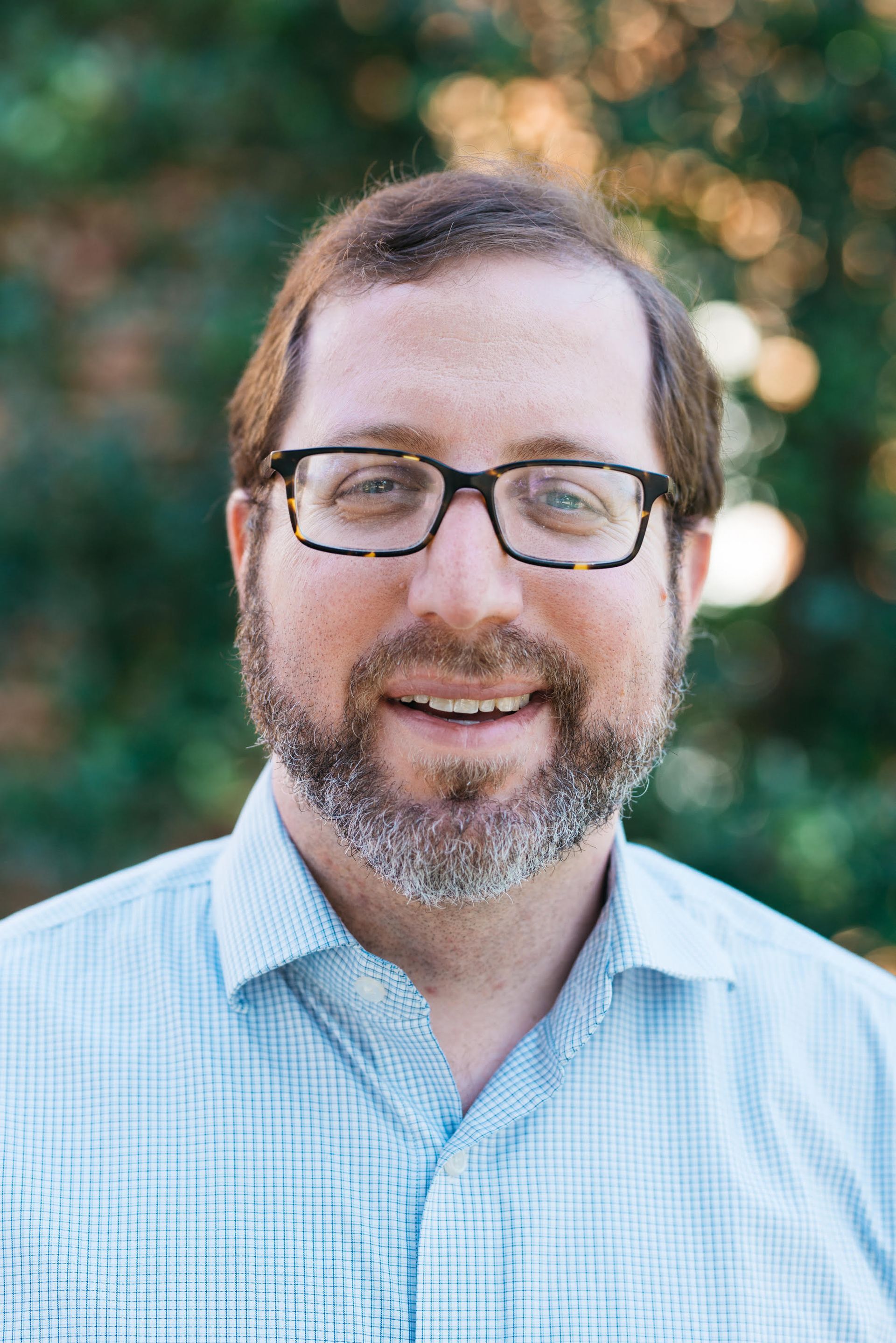 A person with a beard and glasses wearing a light blue button-down shirt, smiling in front of a blurry outdoor background.
