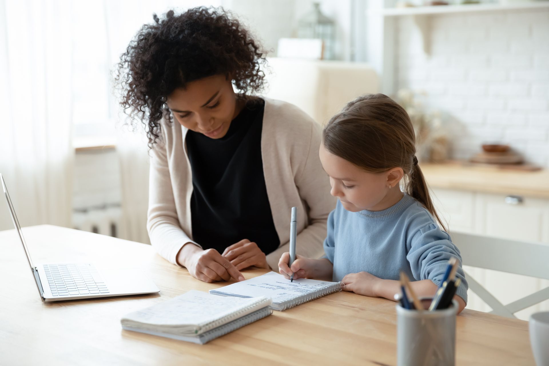 Woman helping a girl write in a notebook at a table, in a kitchen.