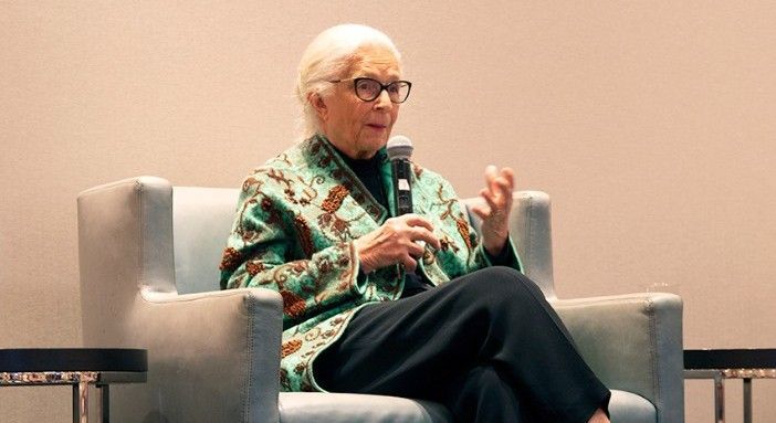 An older woman is sitting in a chair talking into a microphone.
