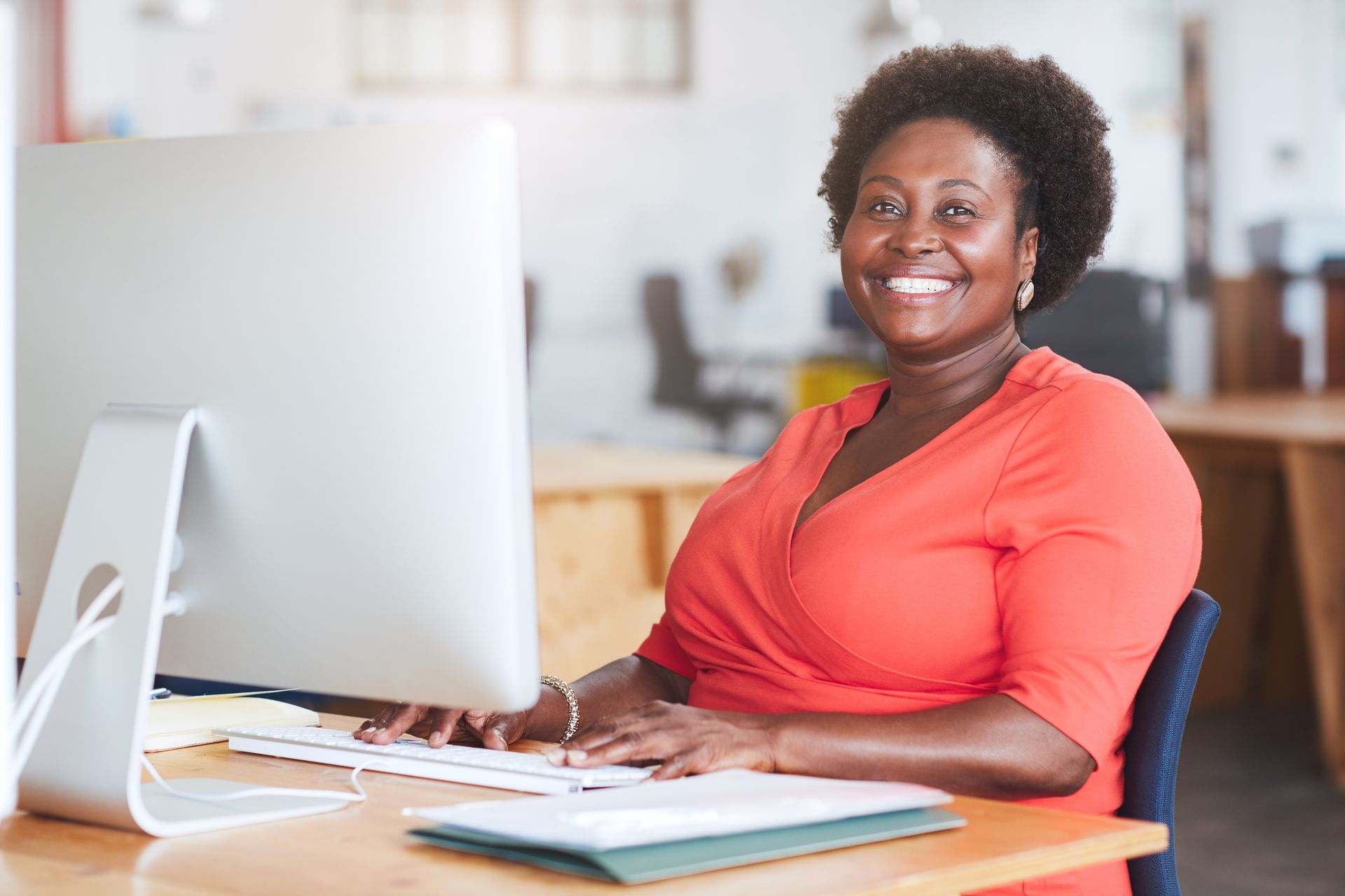 Smiling woman in a coral shirt working at a desk with a computer in a bright office