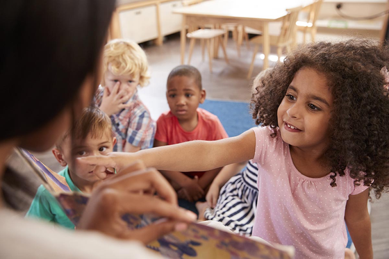 Teacher reads a book to a group of children in a classroom. Girl points at the book, smiling. Other children watch.