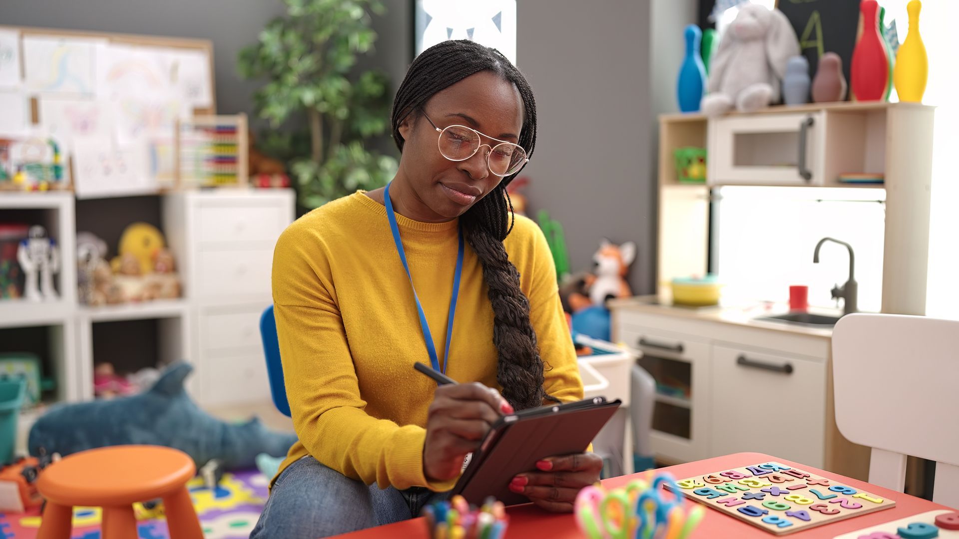 Teacher/Director writing on a tablet in a colorful classroom, with books and supplies around her.
