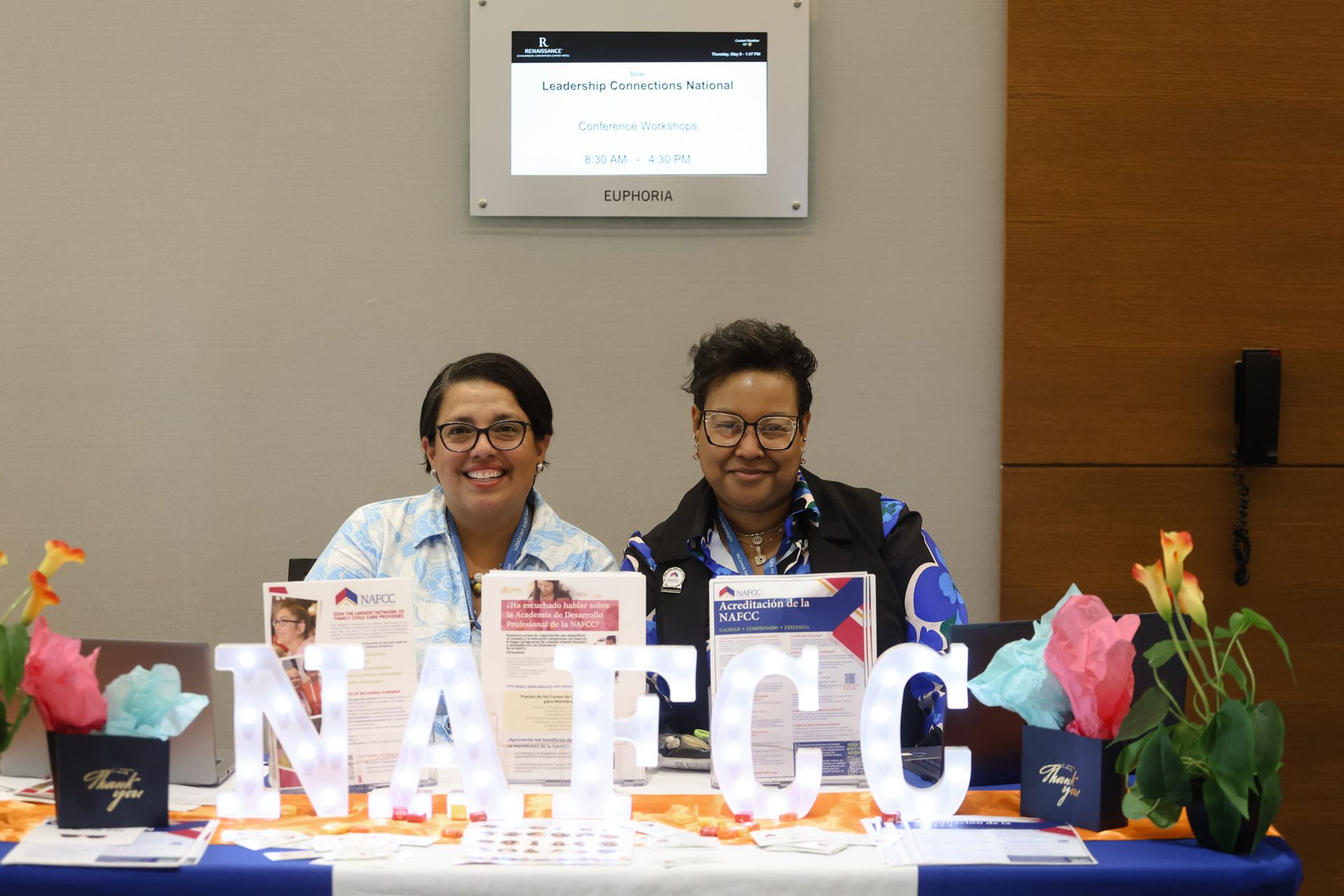 Two people smiling at a table with NATCC letters, promotional materials, and floral decorations.
