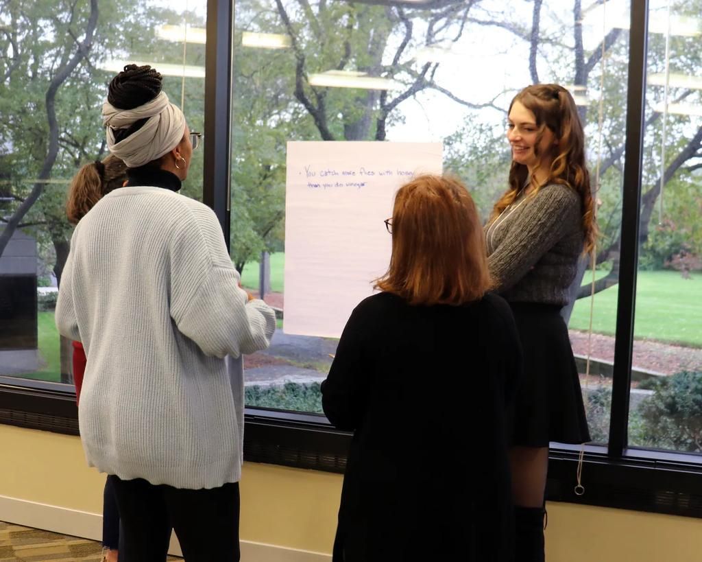 People gathered near a window, viewing a whiteboard with writing; one person gestures. People gathered near a window, viewing a whiteboard with writing; one person gestures.