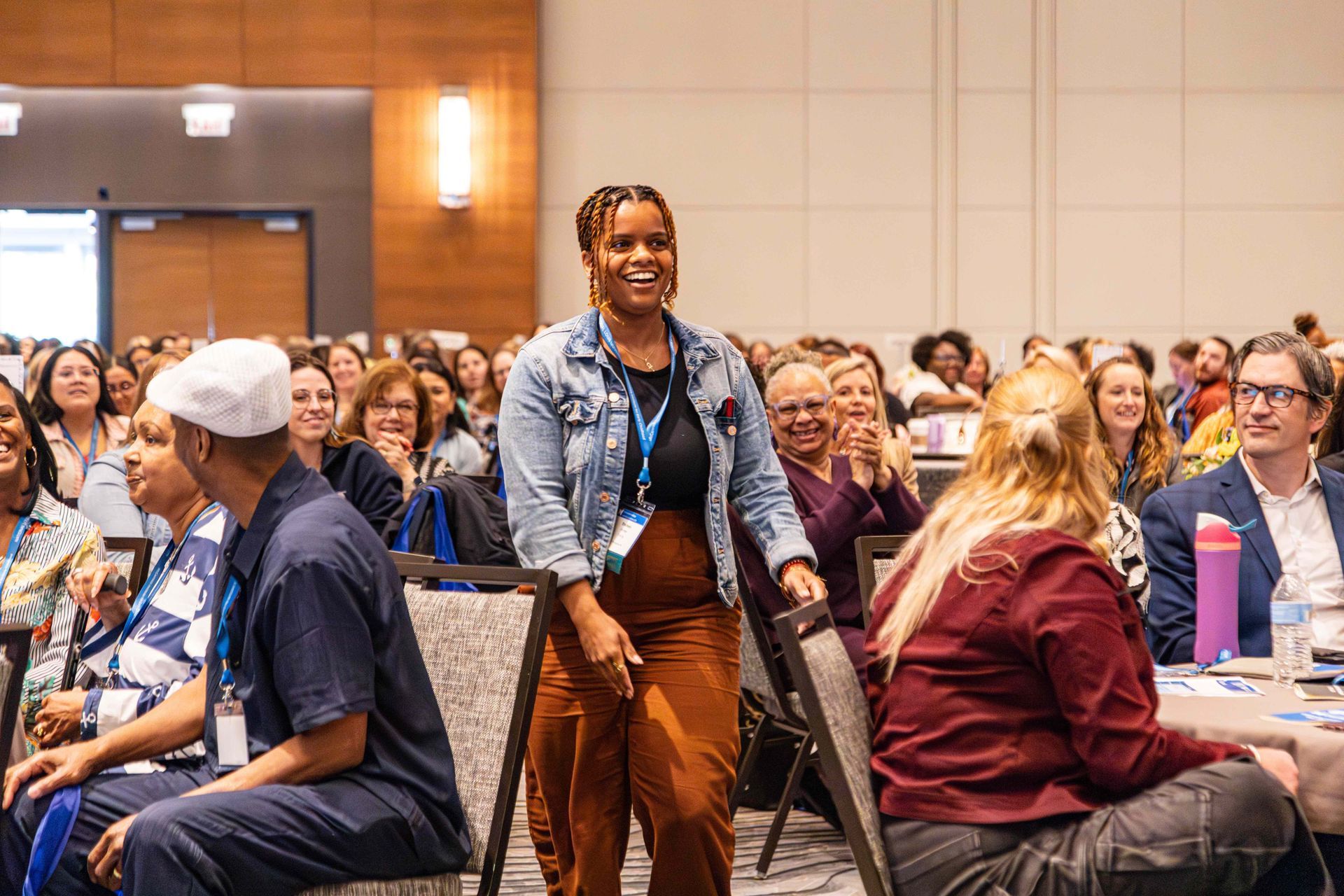 An individual in a denim jacket and brown pants stands smiling in a crowded conference room, surrounded by seated people.