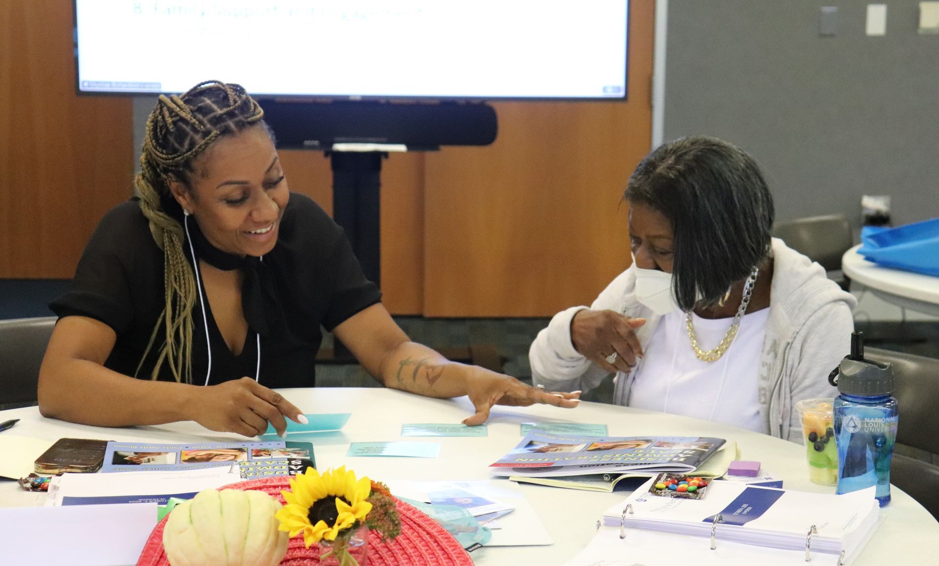 Two people sit at a table drawing and painting with art supplies in a classroom.